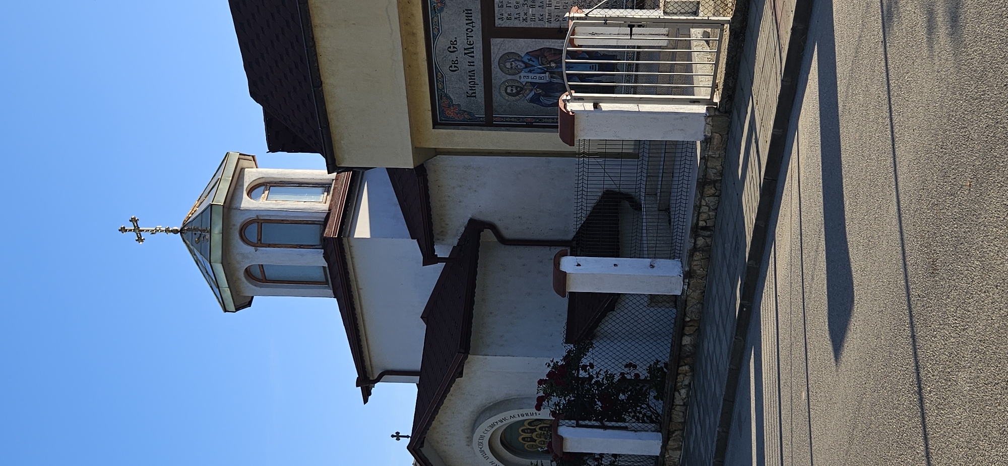 The image shows the exterior of a church, possibly located in Blagoevgrad, Bulgaria. The church has a white facade, a brown roof, and a distinctive tower topped with a cross. One side of the building features an exterior mosaic or panel depicting "Св. Св. Кирил и Методий" (Saints Cyril and Methodius). The adjacent area is enclosed by a metal fence with some vegetation present. A paved road is visible in the foreground, casting long shadows indicating the image was taken during daylight.