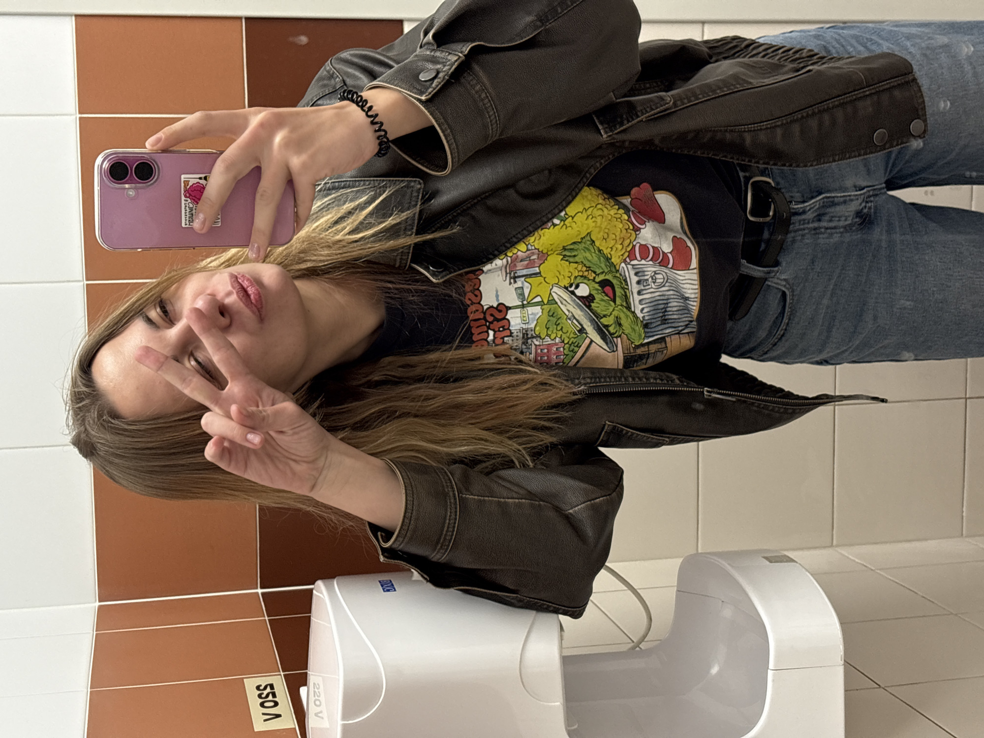 A young woman stands in what appears to be a public restroom, taking a selfie with her phone. She is making a peace sign with her left hand. She is wearing a black t-shirt with a colorful cartoon graphic, a dark brown leather jacket, and blue jeans. A black belt is visible on her jeans. She has a black coiled hair tie on her wrist. Her phone has a pink case with a sticker on the back. Behind her are wall tiles in a two-tone color scheme of white and brown. A white hand dryer is mounted on the wall beneath her. The stickers on the wall read "Voss" and "220 V". The location of the image is unknown and cannot be determined from the image.