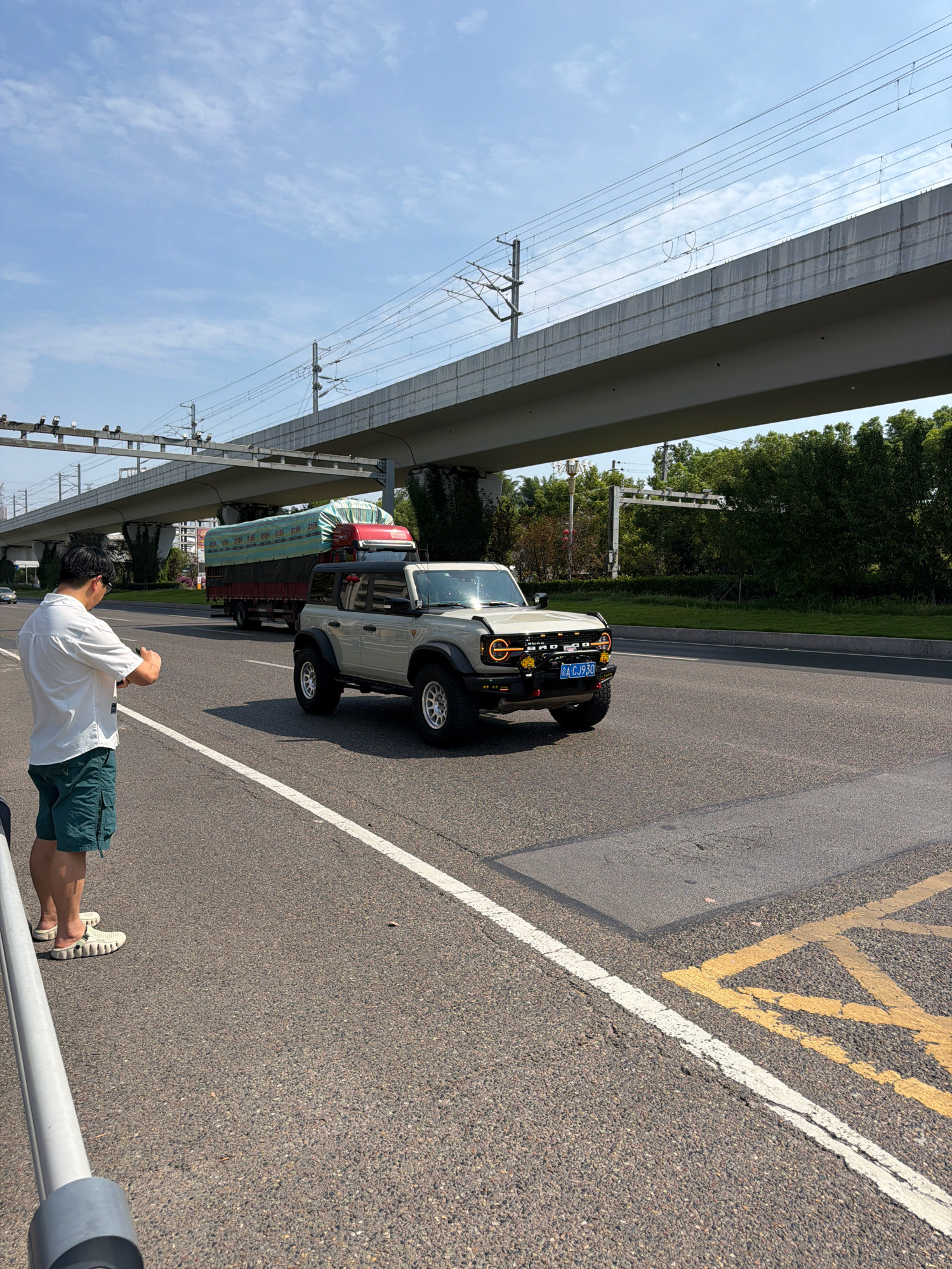 A man stands on the side of a road in Jiangjin, China, holding a mobile phone to take a picture. He is wearing a white short-sleeved shirt and green shorts. A light-colored SUV, possibly a Ford Bronco, is driving past him on the road. The SUV has a blue license plate with Chinese characters. In the background, a large truck with a green tarp covering its load is also on the road. An elevated structure, likely a highway or train track, with overhead wires, runs above the road. Green trees are visible beyond the road. The sky is blue with scattered clouds. The road has white and yellow lane markings.