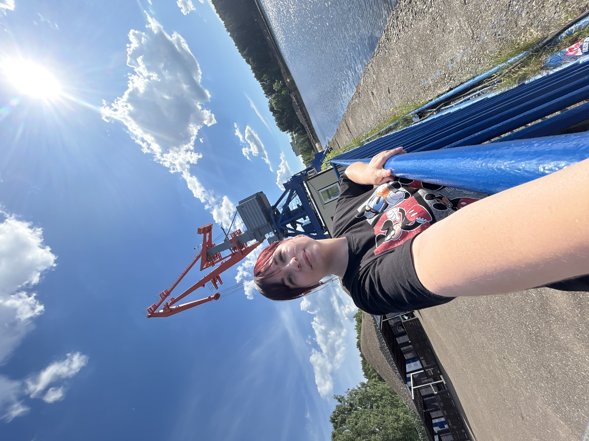 A woman with dark hair and red highlights takes a selfie outdoors on a sunny day. She is wearing a black t-shirt with a Mickey Mouse print. She is standing next to a bright blue railing that overlooks a body of water. In the background, there is a large red crane, some trees, a building with a thatched roof, and a cloudy blue sky. The location is the embankment of the Kama River in Chaikovsky, Russia.