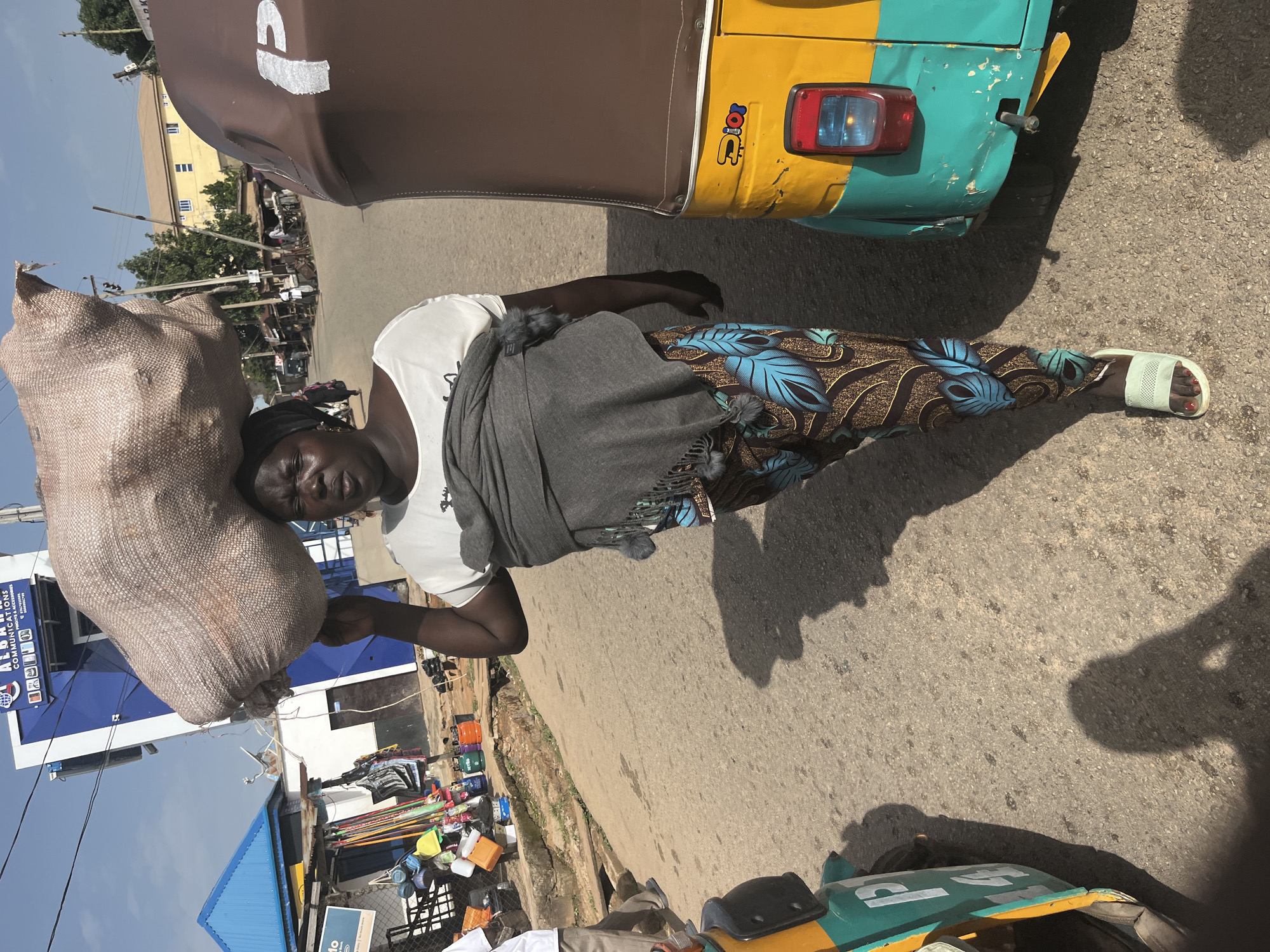 A Nigerian woman, wearing a headscarf, white t-shirt, and patterned pants, carries a large sack of goods on her head as she walks down a street in Gura Top, Nigeria. She is flanked by a yellow and green auto-rickshaw on her right and another auto-rickshaw on her left, with its side visible. In the background, buildings and stalls line the street, and several other people are visible walking or standing. The scene appears to be a bustling market or commercial area on a sunny day.
