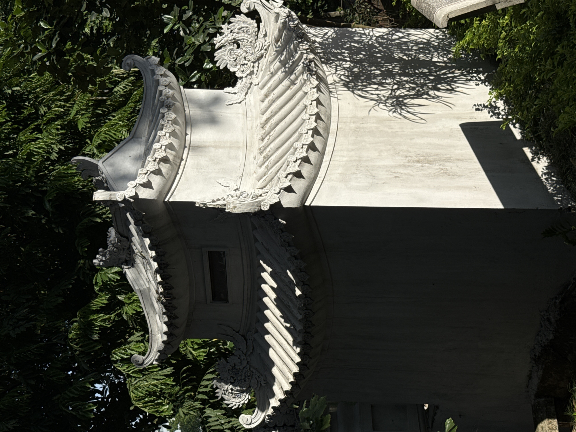 This eye-level shot depicts a section of an ornate, traditional Vietnamese structure, likely a pagoda or temple, located in Hà Nội, Vietnam. The architectural elements include curving roof supports decorated with sculpted details, possibly depicting dragons or floral motifs. The structure is constructed of light-colored stone or concrete. Foliage, including trees and bushes, surrounds the structure, creating a backdrop of deep greens. Shadows cast by the foliage create contrasting light and dark areas on the structure's surface.