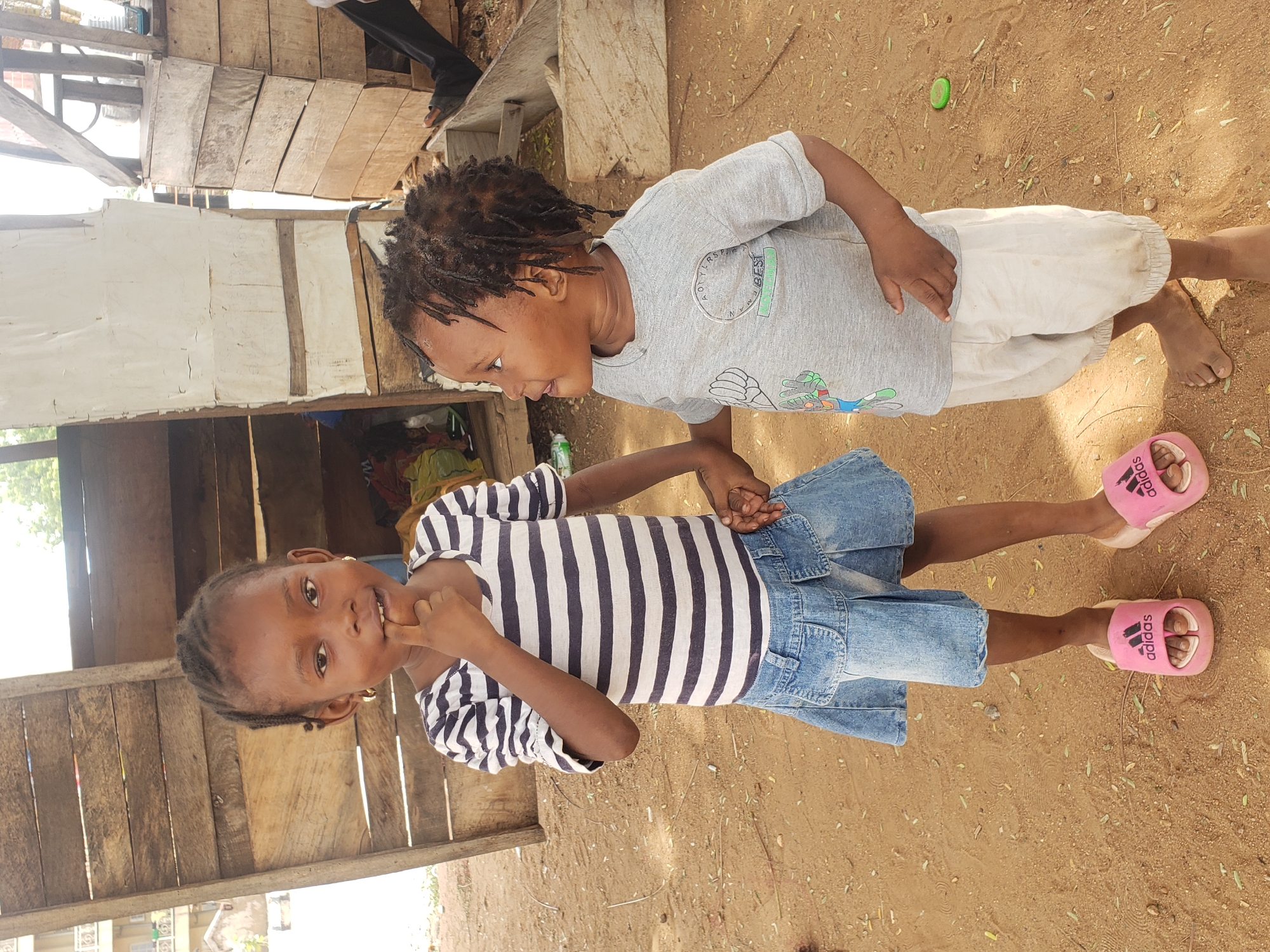 Two children are standing on dirt ground in what appears to be the city of Adankolo, Nigeria. One child is wearing a striped shirt and a blue denim skirt, and pink sandals. The other child is wearing a grey t-shirt and light colored pants. The children are holding hands and looking at each other. A weathered wooden structure is in the background.