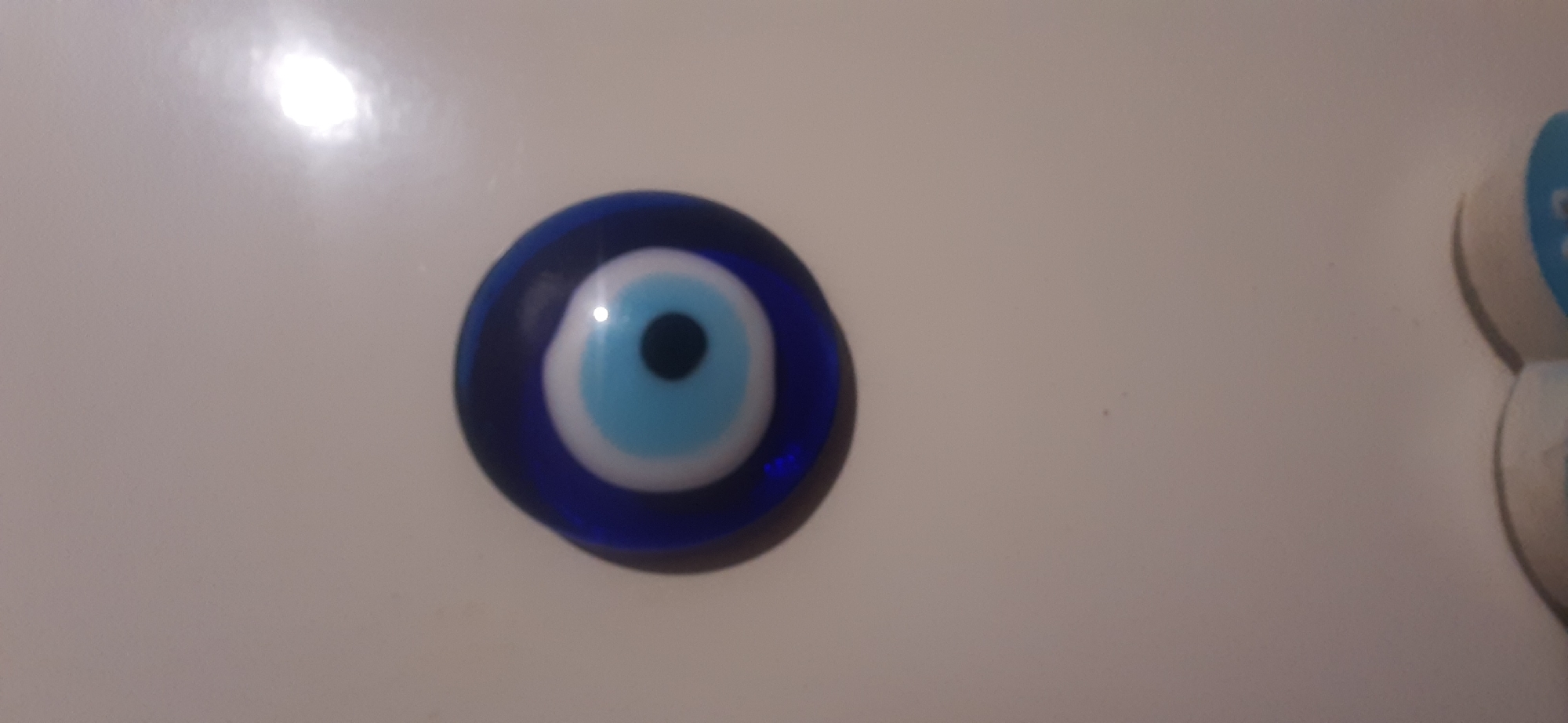 A close-up, top-down shot shows a single, blue evil eye charm on a light-colored, smooth surface. The charm is made of glass and features concentric circles of blue, white, and light blue, with a black pupil at the center. The surface appears to be a countertop or a similar flat area. There are no people or other objects visible in the frame.