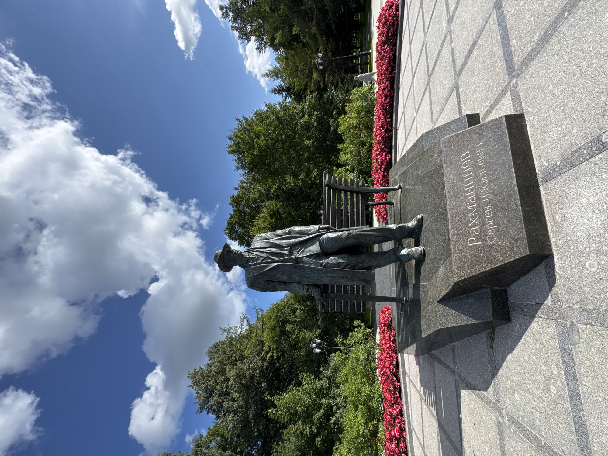 This image shows a bronze statue of a man wearing a hat and a trench coat, standing next to a bench in a park. The statue is located in Veliky Novgorod, Russia. The man in the statue is identified by the inscription on the base as Rachmaninoff, Sergei Vasilievich. The park is filled with lush green trees and a vibrant flowerbed of red flowers. The sky is blue with scattered white clouds. In the background, a person can be seen walking in the park. The statue is placed on a granite pedestal. The ground in front of the statue is paved with granite tiles. The lighting suggests it is daytime.