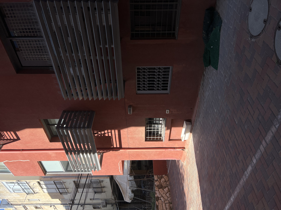 The scene depicts an exterior courtyard or alleyway between two buildings in an urban environment consistent with Tel-Aviv, Israel.
The prominent structure on the right is a multi-story building with a reddish-brown stucco facade. It features two cantilevered balconies on an upper level, both constructed with horizontal gray metal slats. The larger balcony on the right has bird deterrent spikes along its top edge. Several windows are visible: two upper-level windows with dark frames, a lower-level window secured with a white grid, and a ground-level window protected by a closed, collapsible accordion-style security gate. A third ground-level window, partially visible on the far right, has dark security bars. Faint vertical stress cracks are present on the left side of the reddish-brown facade. A small white utility box is mounted below the accordion gate, and a white rectangular planter rests on the ground below the grid-barred window.
The ground is paved with reddish-brown interlocking bricks arranged in a herringbone pattern. A faded white line traverses the brickwork in the lower-left quadrant. Two circular metal manhole covers are visible in the lower right. A dark green mat or tarp, partially rolled, lies against the base of the red building in the lower right corner.
In the background, to the left, stands a taller, lighter-colored (beige/off-white) building. This structure appears older, featuring multiple windows and balconies with dark railings. Overhead, several dark utility cables stretch horizontally. An external air conditioning unit is mounted on this building's facade. Below, a light-colored canopy or awning extends over a fenced area. A low retaining wall constructed from stacked, light-colored stones borders the left edge of the paved area, with some sparse vegetation behind it.
Strong sunlight illuminates the scene, casting sharp, defined shadows from the balconies and portions of the red building onto its facade and the paved ground. No individuals are present in the image, and no active interactions or discernible movements are depicted.