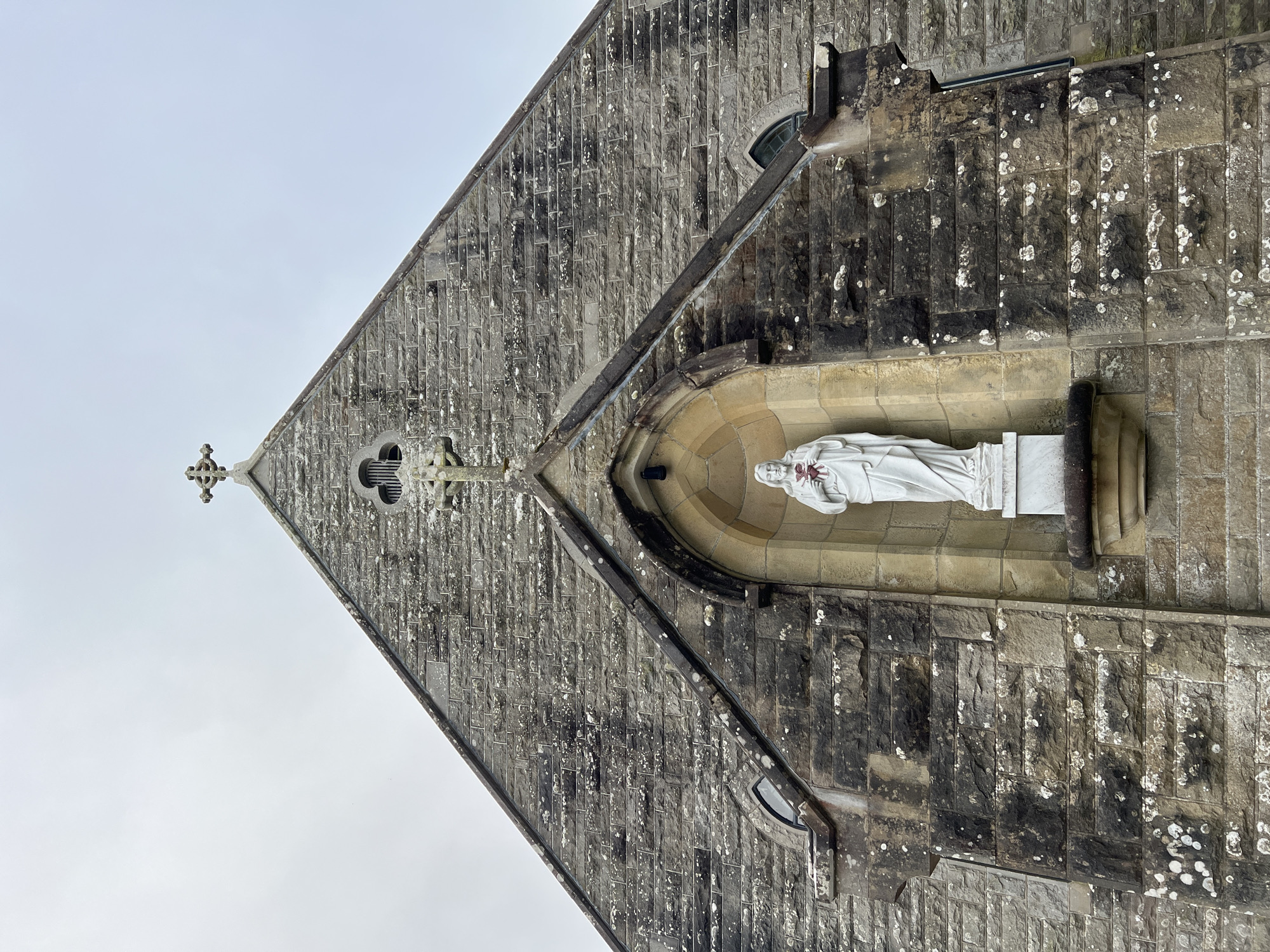 The image shows a low-angle, full-frame shot of the facade of a stone church in Mountcharles, Ireland. A white statue of Jesus, holding a red heart, is positioned in an arched niche in the center of the building. The church's gable is topped with a stone cross. The stone blocks making up the facade are weathered and discolored, with patches of lichen. Small arched windows are visible on either side of the niche. The sky is overcast.