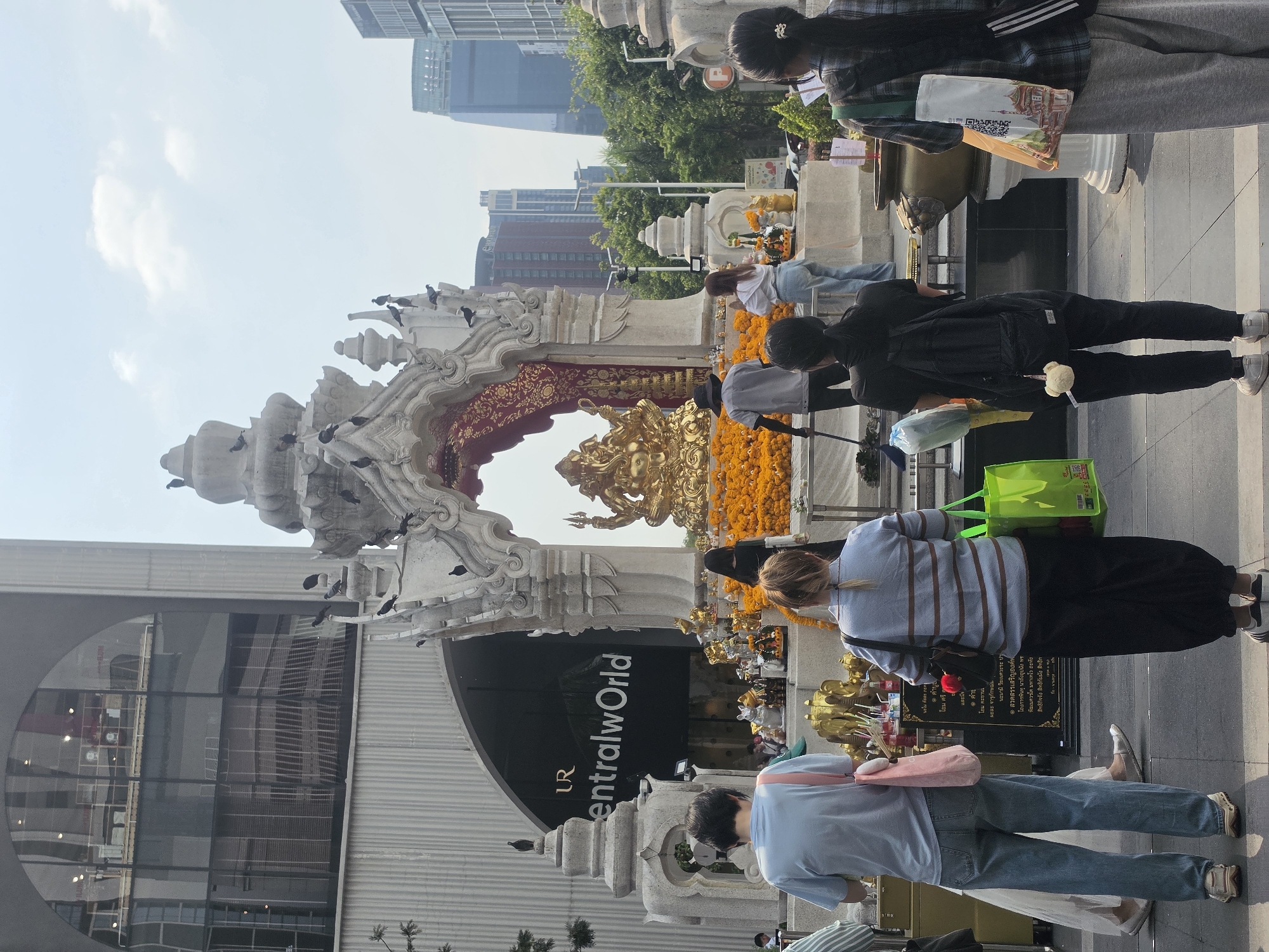 The image shows a religious shrine in Bangkok, Thailand, located near the CentralWorld shopping mall. The shrine features a golden statue under an arch, adorned with orange garlands. People are seen making offerings and praying at the shrine. The environment includes city buildings and a clear sky. Pigeons are perched on the structure of the shrine. The area appears to be a public space with pedestrian traffic.
