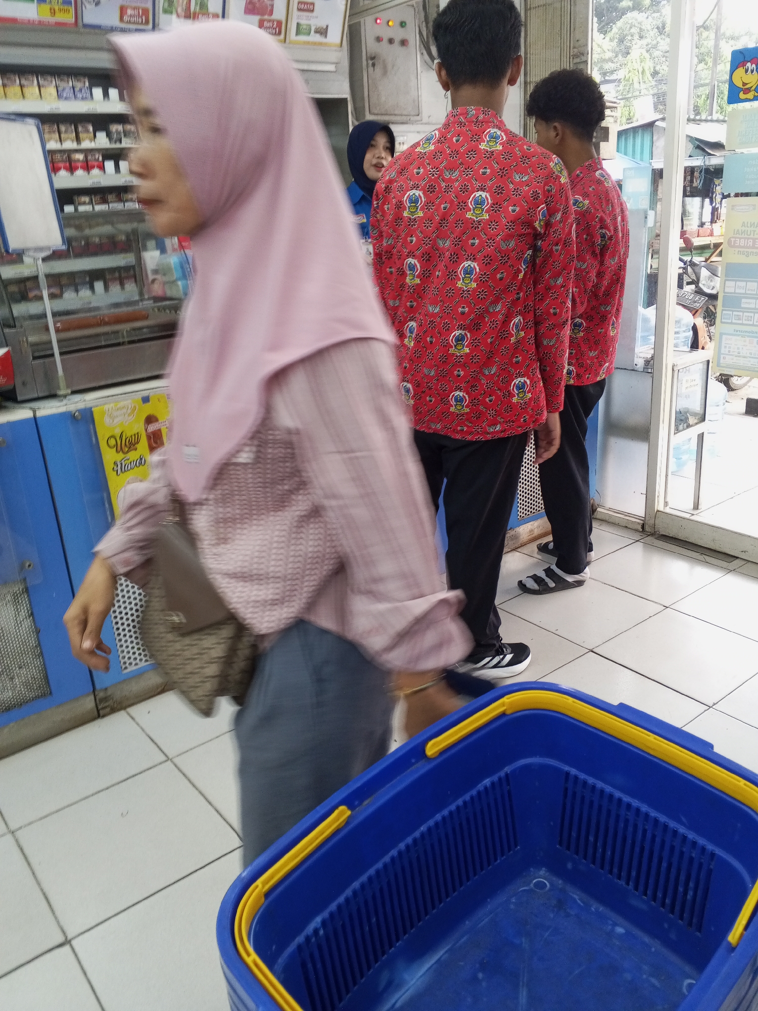 In Ciruas, Indonesia, an outdoor convenience store scene unfolds. A woman wearing a pink hijab and a plaid shirt walks past a display of cigarettes, a brown handbag slung across her body. To her right, two young men in matching red shirts with intricate patterns stand near the entrance. One man wears black pants and black and white athletic shoes, while the other has black pants and black sandals. In the foreground, a bright blue shopping basket with yellow handles sits on a tiled floor. The store's interior, visible through the open glass doors, shows shelves stocked with products and signs advertising various goods. The environment appears to be a busy commercial area with a focus on retail.