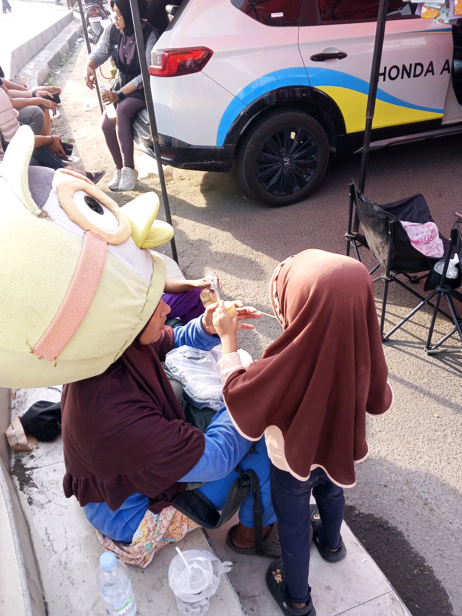 In Serang, Indonesia, a young person wearing a large owl-themed costume headpiece sits on a concrete ledge next to a white Honda car with blue and yellow decals. The costume head is pale yellow with orange and pink accents around the eyes and ears. The person is dressed in a dark brown long-sleeved shirt and bright blue pants. Another young person, wearing a brown hijab and a brown long-sleeved shirt, stands next to the first, holding a stick with a yellow object on the end, possibly food or a toy. They appear to be interacting. In the background, a woman wearing a hijab and glasses sits on a black folding chair next to the car, holding a plastic bottle of water. The car has "HONDA A" visible on its side. The ground is paved with asphalt. Other people are partially visible in the background, along with some parked motorcycles. The scene appears to be outdoors during daylight hours.