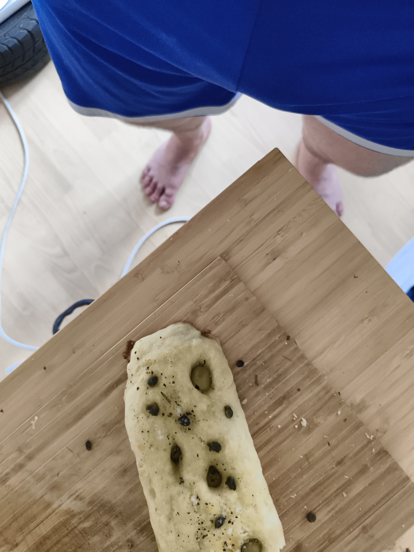A high-angle shot shows a section of focaccia bread on a light-colored wooden board, likely a cutting board. The focaccia is topped with green olives and a sprinkling of what appears to be salt and herbs. Adjacent to the cutting board, the viewer sees a portion of a person's legs, clad in blue athletic shorts with a light-colored trim. The person is standing barefoot on a light-colored wooden floor. A black tire can be seen in the background of the upper left corner of the image. A white cable is trailing in the background as well. It is impossible to determine a specific location within the city of Diepholz, Germany from the image alone.