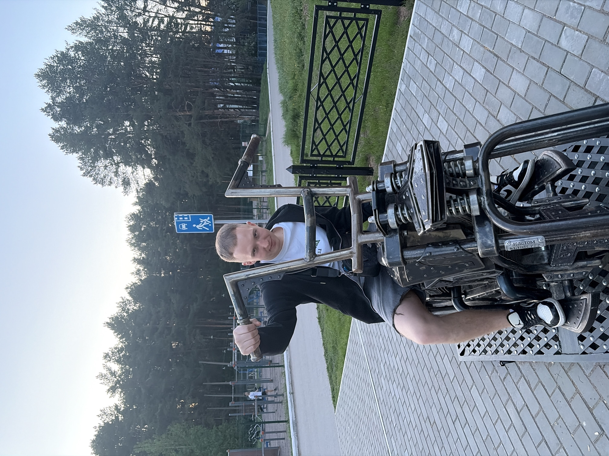 A man is seated on a metal outdoor exercise machine, likely in Chaikovsky, Russia. He is wearing a white t-shirt, black jacket, and grey shorts. The machine has footrests and handles. The location appears to be a park or recreational area with trees, a metal fence, and a paved walkway. A blue sign with a bus and pedestrian symbol is visible in the background. Other people are present, visible in the distance engaging in outdoor fitness activities.