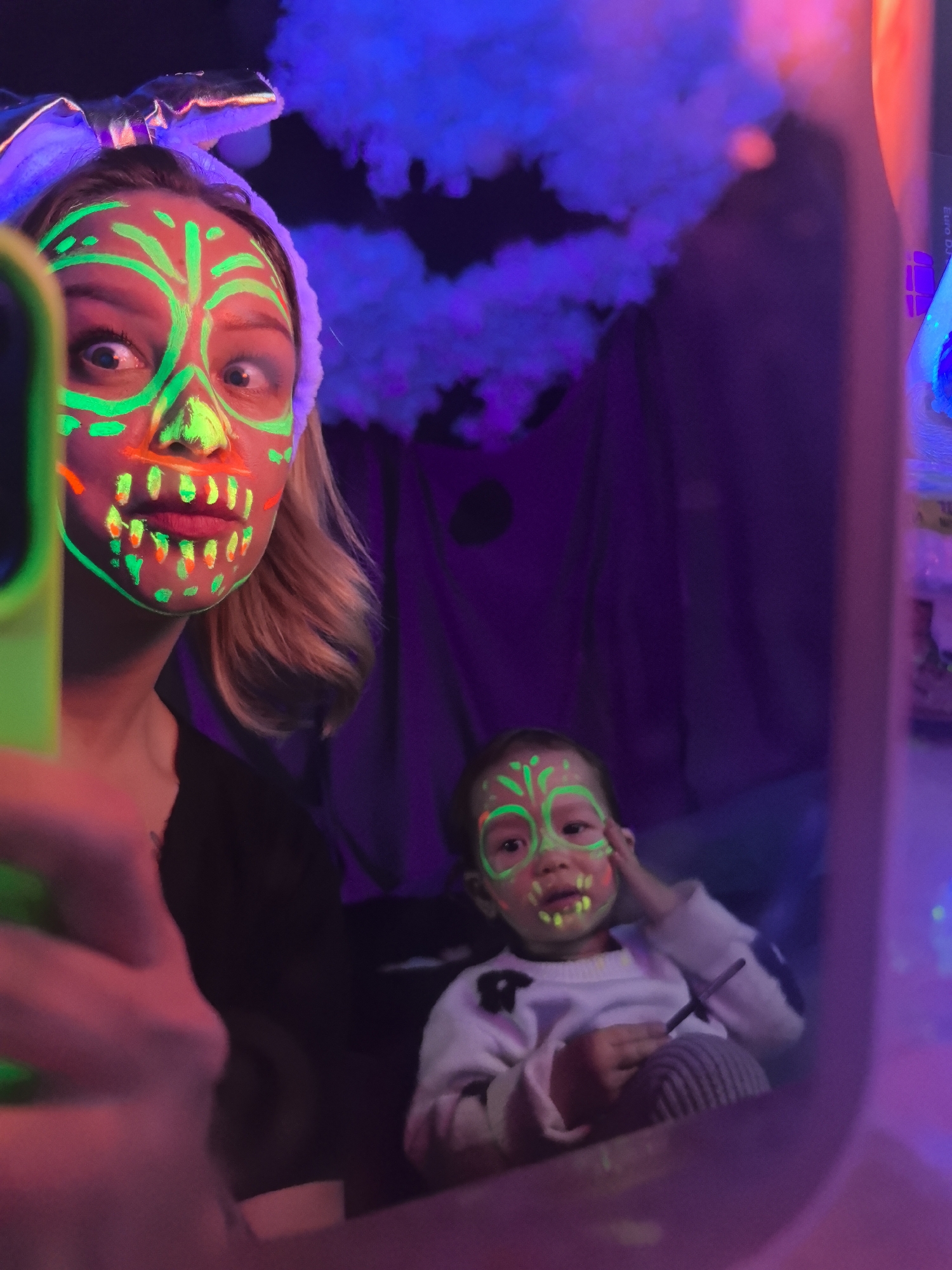 A woman and a toddler are seen in a mirror selfie. Both have green neon face paint; the woman has rabbit ears. The woman is holding a cell phone to take the photo. The toddler sits in the background in front of a purple curtain with blue lights. The location is Noviy, Russia.
