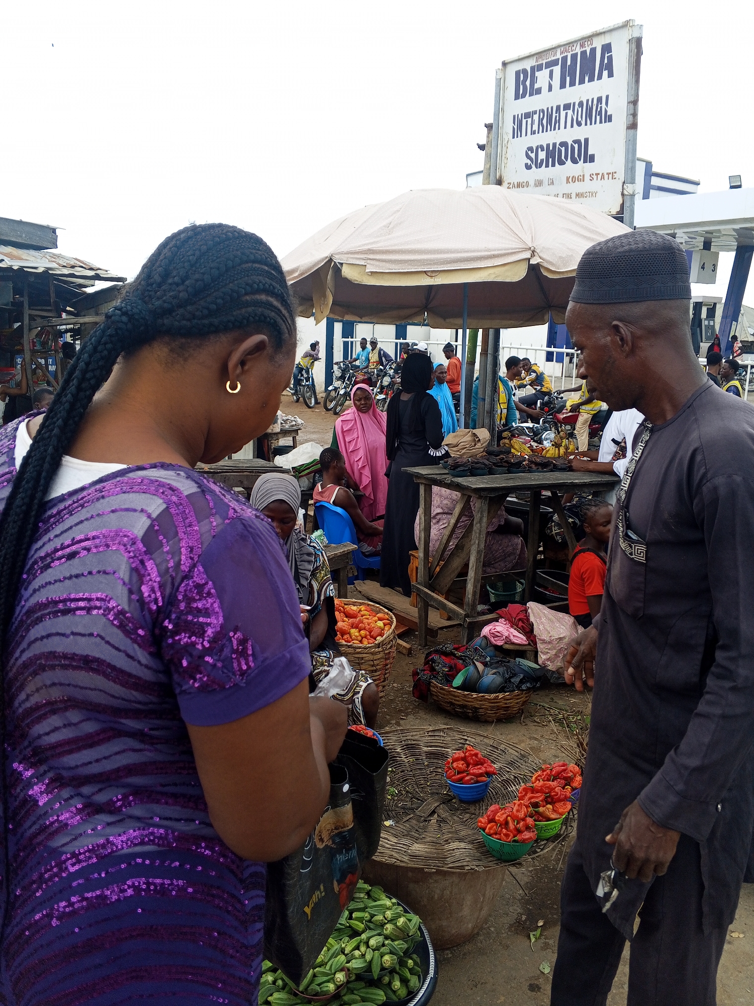 The image depicts a bustling market scene in Zango-ndaji, Nigeria, with a clear view of a "BETHMA INTERNATIONAL SCHOOL" sign in the background. In the foreground, a woman with braided hair, wearing a purple sequined top, is seen from behind, interacting with a market stall. She is holding a black bag with colorful graphics. To her right, a man wearing a dark cap and a dark outfit stands facing away from the camera, appearing to be involved in the market activity.
The market stall features an array of fresh produce. In the foreground, a basket overflowing with green okra is visible. Next to it, three bowls contain vibrant red peppers, some of which are conical in shape. A woven mat forms the surface of the stall, on which other items are displayed, including baskets of tomatoes. Further back, there are wooden tables laden with more goods, possibly food items, and shoppers are visible in the background, some on motorcycles. The overall environment appears to be an open-air market under a large, cream-colored umbrella, with makeshift stalls and a busy atmosphere. The sky is overcast.