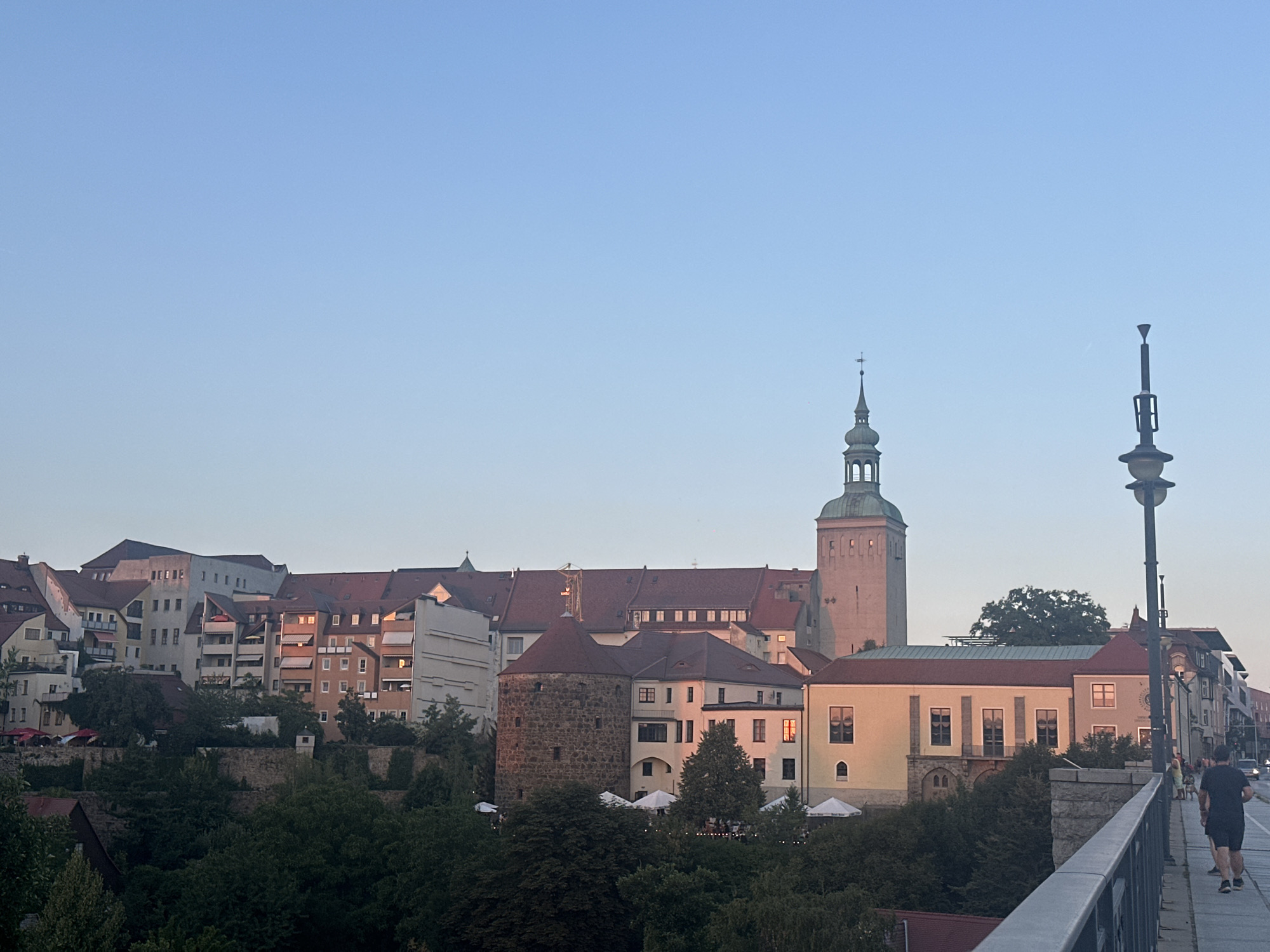 The image depicts a sunset view over the city of Bautzen, Germany. In the foreground, a stone bridge with a metal railing spans across the frame. A single figure, dressed in dark athletic wear, is captured in mid-stride, jogging away from the camera on the bridge's pedestrian path. Beyond the bridge, the cityscape unfolds. Several buildings with red-tiled roofs are visible, interspersed with trees. A prominent round stone tower with a conical roof stands out, and to its right, a taller, slender tower with a green, domed roof and a cross atop it pierces the pale blue sky. White umbrellas suggest outdoor seating areas amidst the greenery. The setting sun casts a warm glow on some of the buildings, highlighting their architectural details. The sky is clear and transitions from a lighter shade near the horizon to a deeper blue overhead.