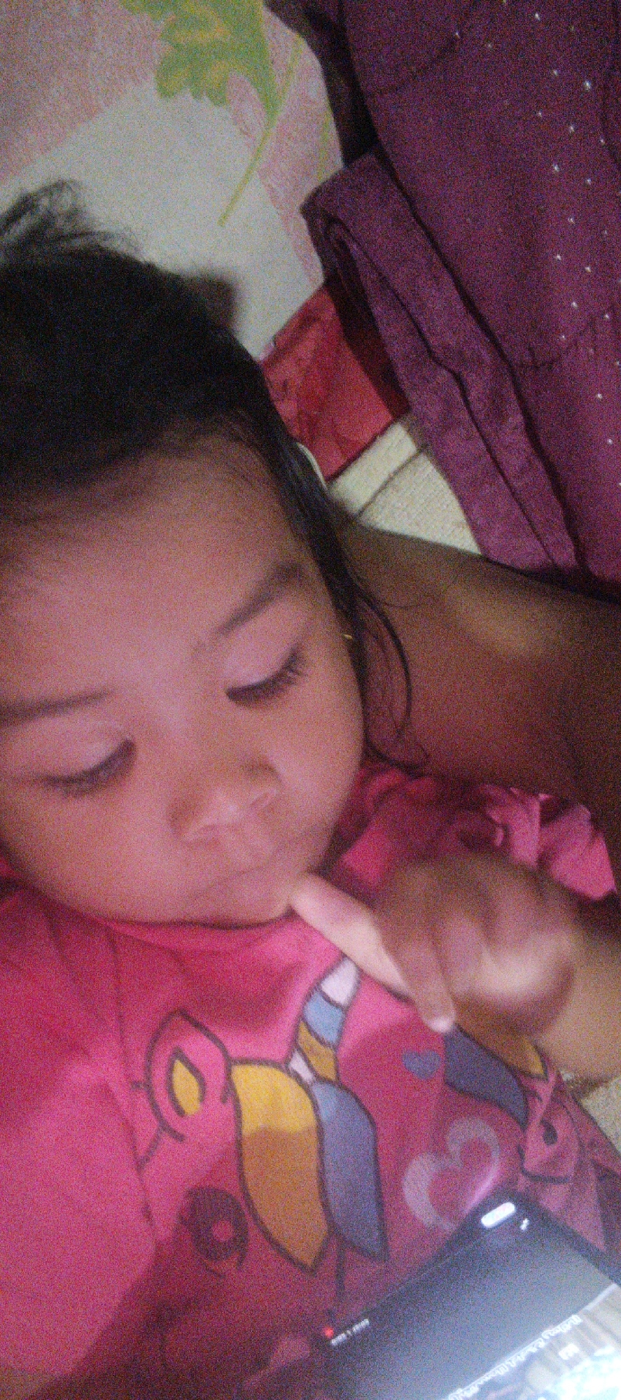 A close-up, slightly grainy, eye-level shot shows a young child wearing a pink shirt with a cartoon design. The child is lying down, with their head turned to the left and their finger touching their chin. A dark blanket is visible behind and to the right of the child, with a lighter patterned blanket in the background. The scene appears to be indoors, possibly in a bedroom. The location in Cinta Dame, Indonesia, cannot be determined from the image.
