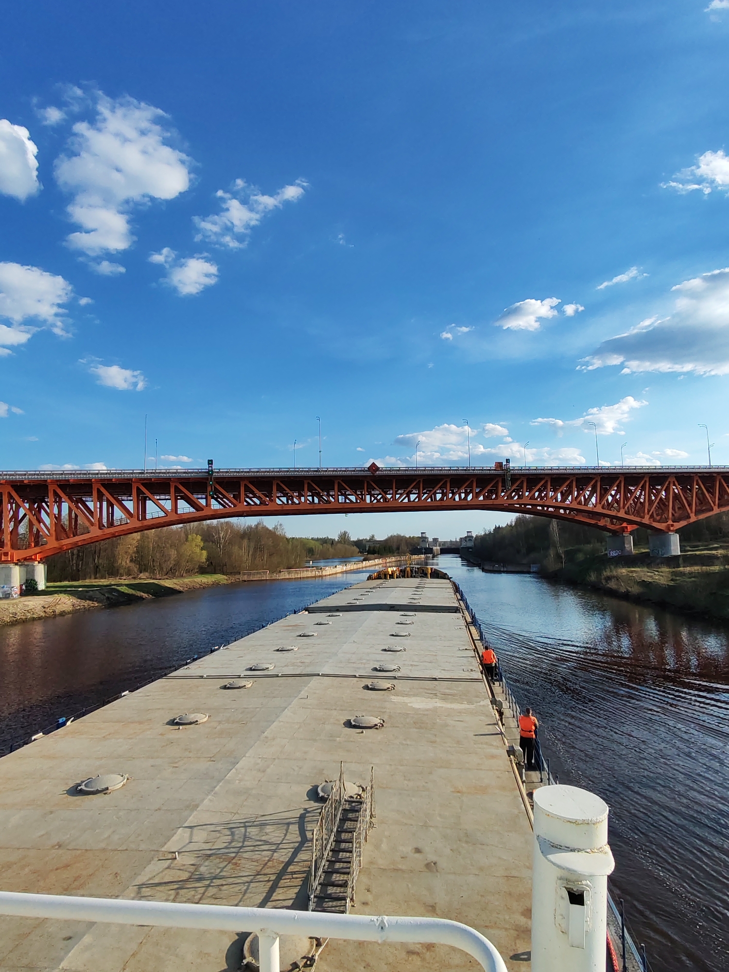 The image shows a barge, presumably on the Volga River near Ples, Russia, approaching an orange bridge. Two people wearing orange safety vests stand on the edge of the barge. The barge is wide and flat, with numerous round access covers. The river reflects the trees and sky. Above, a blue sky is dotted with white clouds. The bridge is an orange, steel structure with a truss design and appears to be carrying vehicle traffic. Trees line the riverbanks on either side. The lighting suggests it's a sunny day.