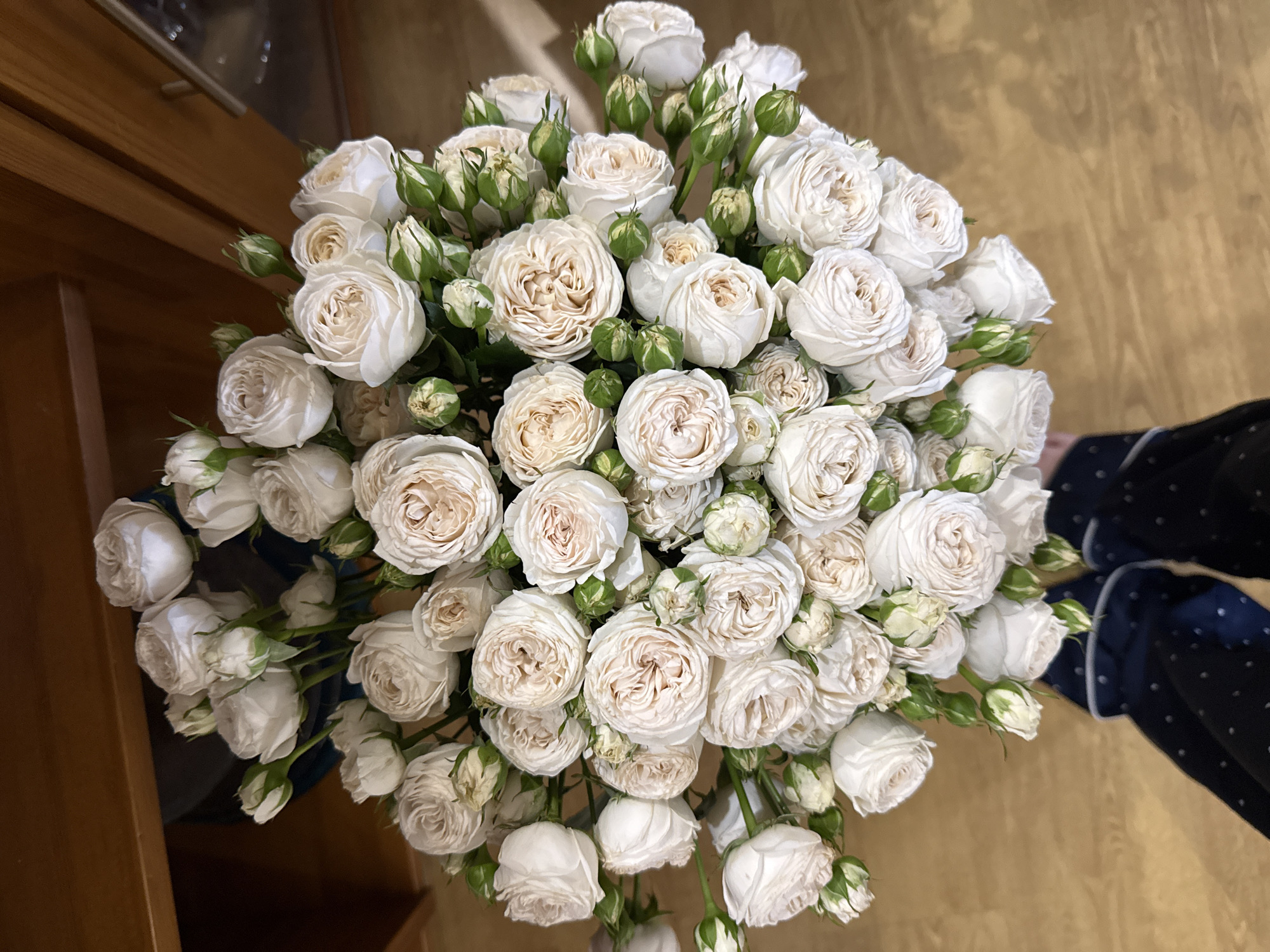 A high-angle, eye-level shot depicts a dense bouquet of white roses with green buds and stems. The roses are of a variety with many petals, and some have a hint of peach or light beige in the center. The background is a blurred mix of wooden surfaces, possibly a cabinet or countertop, and a portion of a person's dark blue patterned pajamas. The location is not identifiable as Krasnoyarsk, Russia from the visual information available. There are no people fully visible in the scene, only a portion of a person's clothing. There is no action apparent, merely the display of a bouquet.