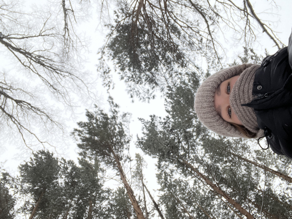 A person wearing a neutral-colored knitted hat and dark jacket looks directly down at the camera. The shot is angled upwards toward the sky, which appears overcast. Surrounding the person are tall pine trees covered in snow or frost. Some bare deciduous trees are also visible in the background, creating a stark contrast against the white sky.