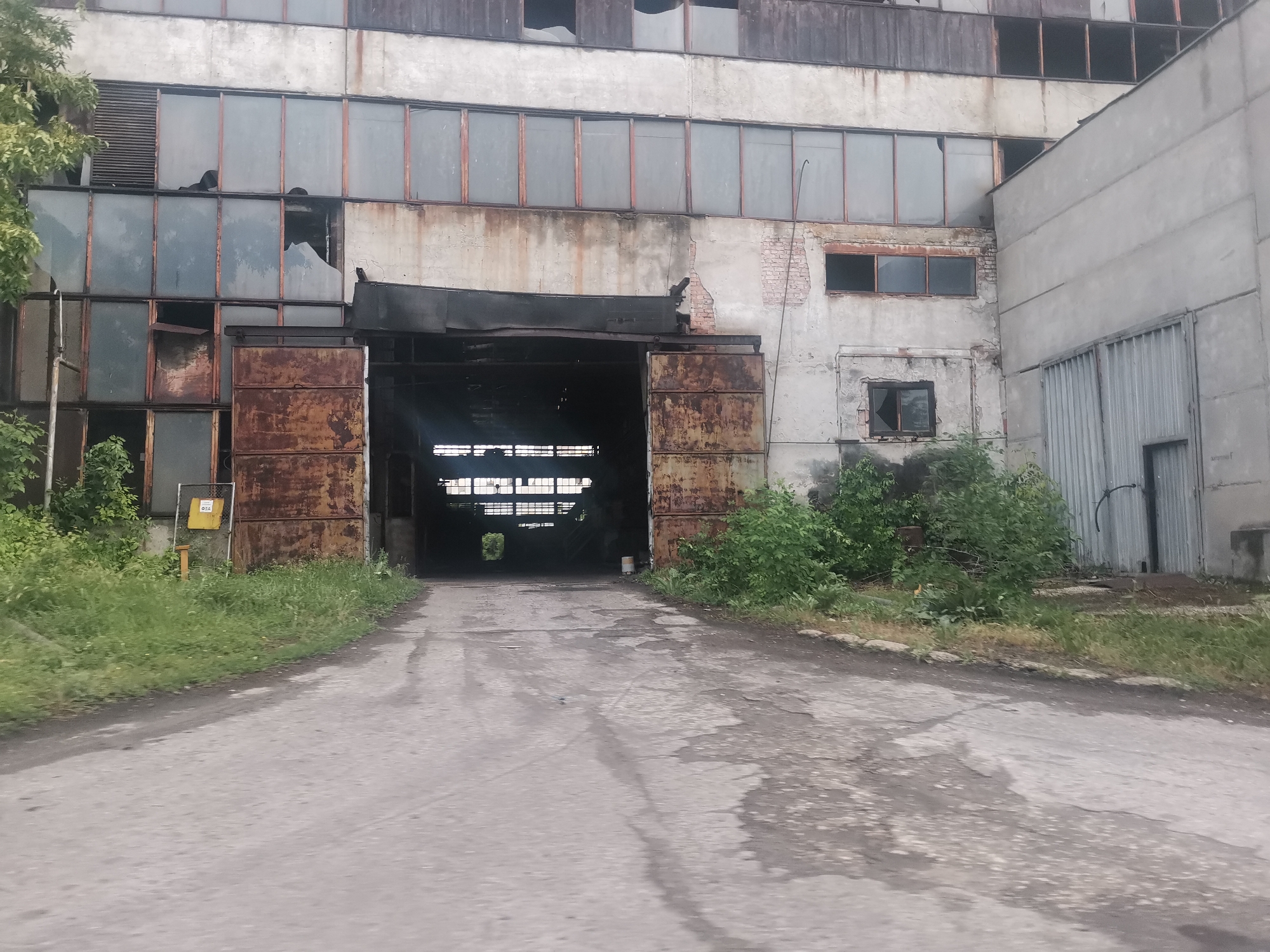The image depicts an abandoned, multi-story industrial building or factory in Zhivkovo, Bulgaria, viewed from a paved driveway. The scene portrays a state of significant disrepair and neglect.
The building's facade is primarily concrete, with numerous windows arranged in rows. Many of these windows are broken, cracked, or missing panes, revealing dark interior spaces. Some window frames show visible rust and decay. Patches of exposed brickwork are visible, particularly near the main entrance.
A large, dark archway forms the central entrance to the building, flanked by heavily rusted metal gates or doors that appear inoperative. Above this entrance, a dark, overhanging structure shows signs of partial collapse. Through the archway, the interior is dimly lit, with grid-like light sources visible further inside, suggesting additional windows or skylights.
To the right of the main entrance, another section of the building features fewer windows and includes a grey corrugated metal gate or door. Lush, overgrown vegetation, comprising tall grass and various bushes, frames the building's base and edges of the driveway, emphasizing the abandonment. On the left side, a small yellow sign displaying the Cyrillic text "ФБА" is partially visible amongst the greenery.
The paved driveway in the foreground is cracked, worn, and appears wet or stained in places, indicating weathering and lack of maintenance. The lighting suggests daytime under an overcast sky, as there are no harsh shadows. No people are present, and no specific activities are taking place, reinforcing the impression of a deserted industrial site. The overall mood is one of decay and desolation.
