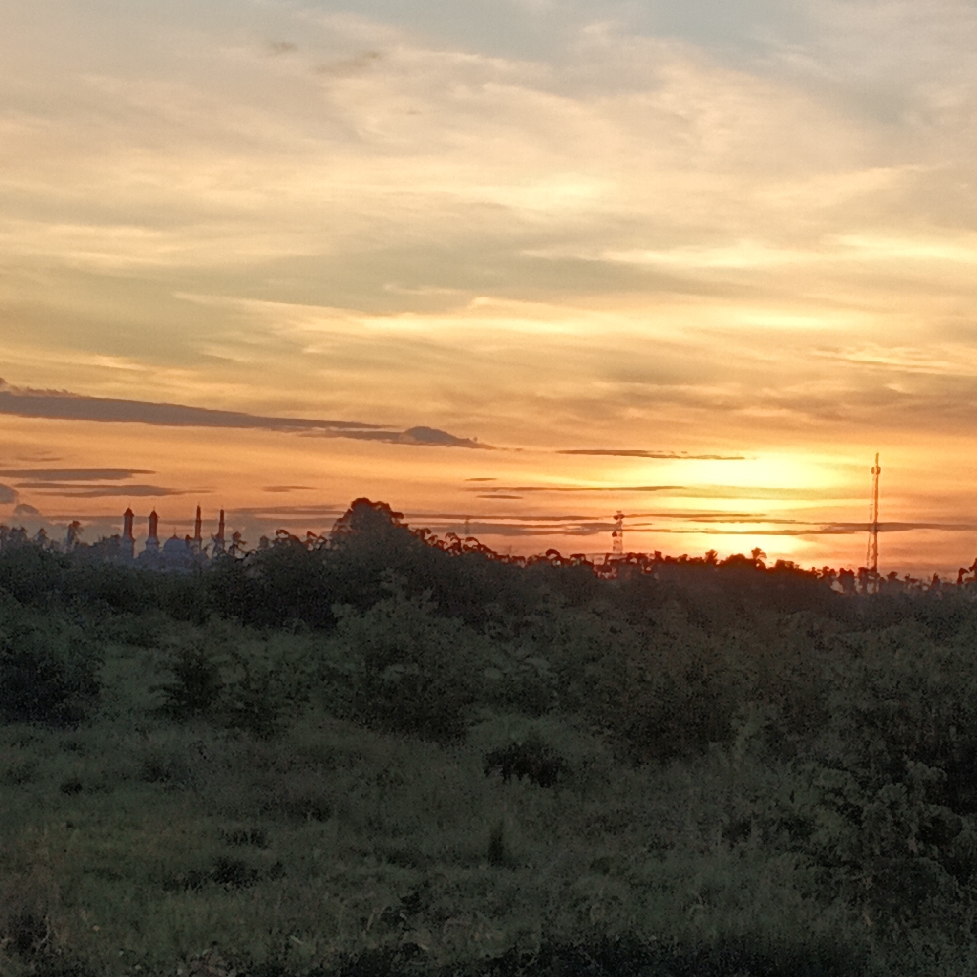 The image depicts a sunset over a landscape in Dayah Kruet, Indonesia. In the foreground, a dense area of dark green foliage is visible, obscuring the ground. Beyond the foliage, a silhouetted cityscape emerges, featuring several distinct architectural structures that appear to be minarets and domes, suggesting a mosque or a cluster of religious buildings. To the right, a tall, thin telecommunications tower stands prominently against the sky. The sky itself is a spectacle of sunset colors, with horizontal bands of orange, yellow, and lighter blue clouds stretching across the frame. The sun is setting in the distance, casting a warm glow that illuminates the clouds. The overall impression is a serene, twilight scene of a rural or semi-urban area with a noticeable religious presence.