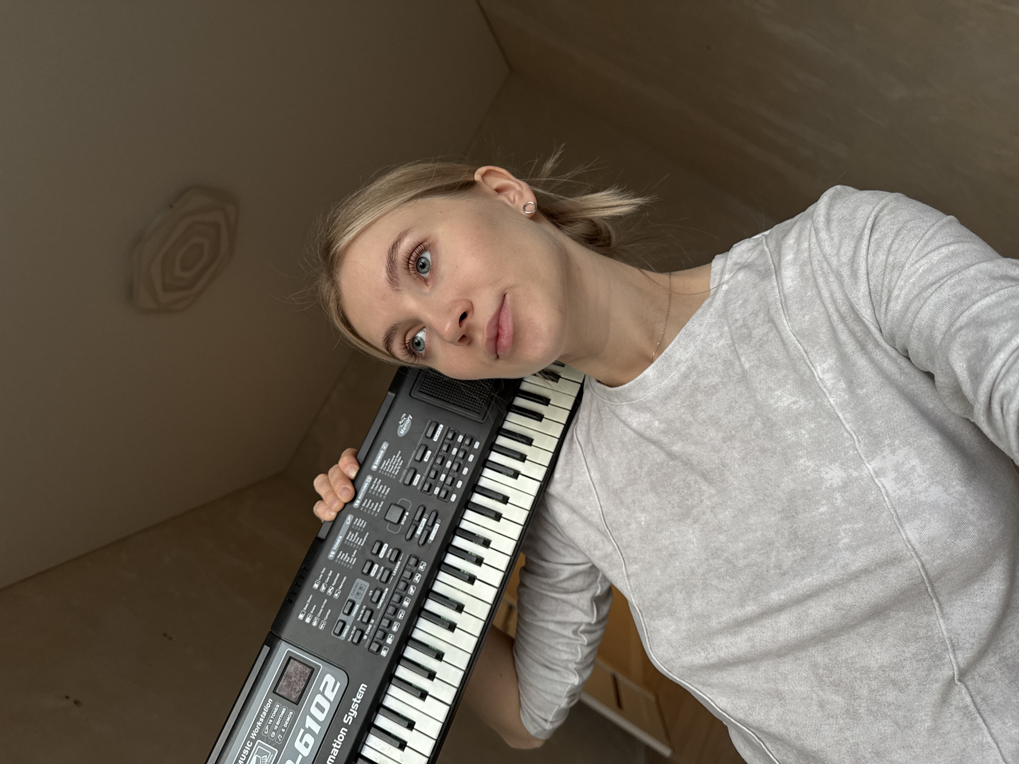 A fair-skinned woman with blonde hair partially pulled back is holding a black electronic keyboard against her cheek. The keyboard has white and black keys and various buttons and a small screen. The model number "SK-6102" is visible on the keyboard. She is wearing a light grey, long-sleeved shirt and a small silver necklace. The background consists of off-white walls and ceiling with minimal features. The room appears to be unfinished or sparsely decorated. There is no evidence to suggest the location is Izhevsk, Russia.