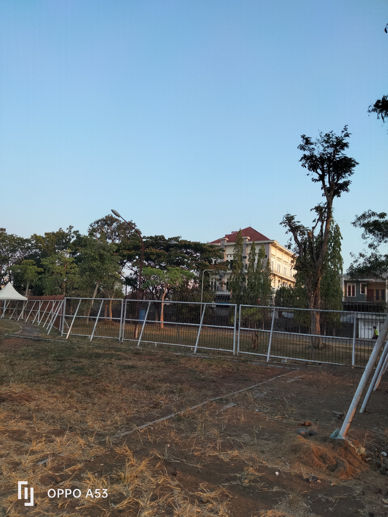 The image captures an outdoor scene under a clear blue sky. In the foreground, dry, light brown grass covers the ground, interspersed with patches of bare earth. A white metal fence with diagonal supports runs across the mid-ground, separating the grassy area from a background of trees and buildings. Behind the fence, several trees with dense green foliage are visible, some with bare branches reaching towards the sky. A multi-story building with a red-tiled roof and white walls stands behind the trees, appearing to be a hotel or similar establishment. The building has numerous windows and architectural details. Further to the right, another smaller building with a darker facade is partially visible. The overall setting appears to be an open area, possibly a park or event space, with urban structures in the distance. The image also displays a watermark in the bottom left corner indicating "OPPO A53".