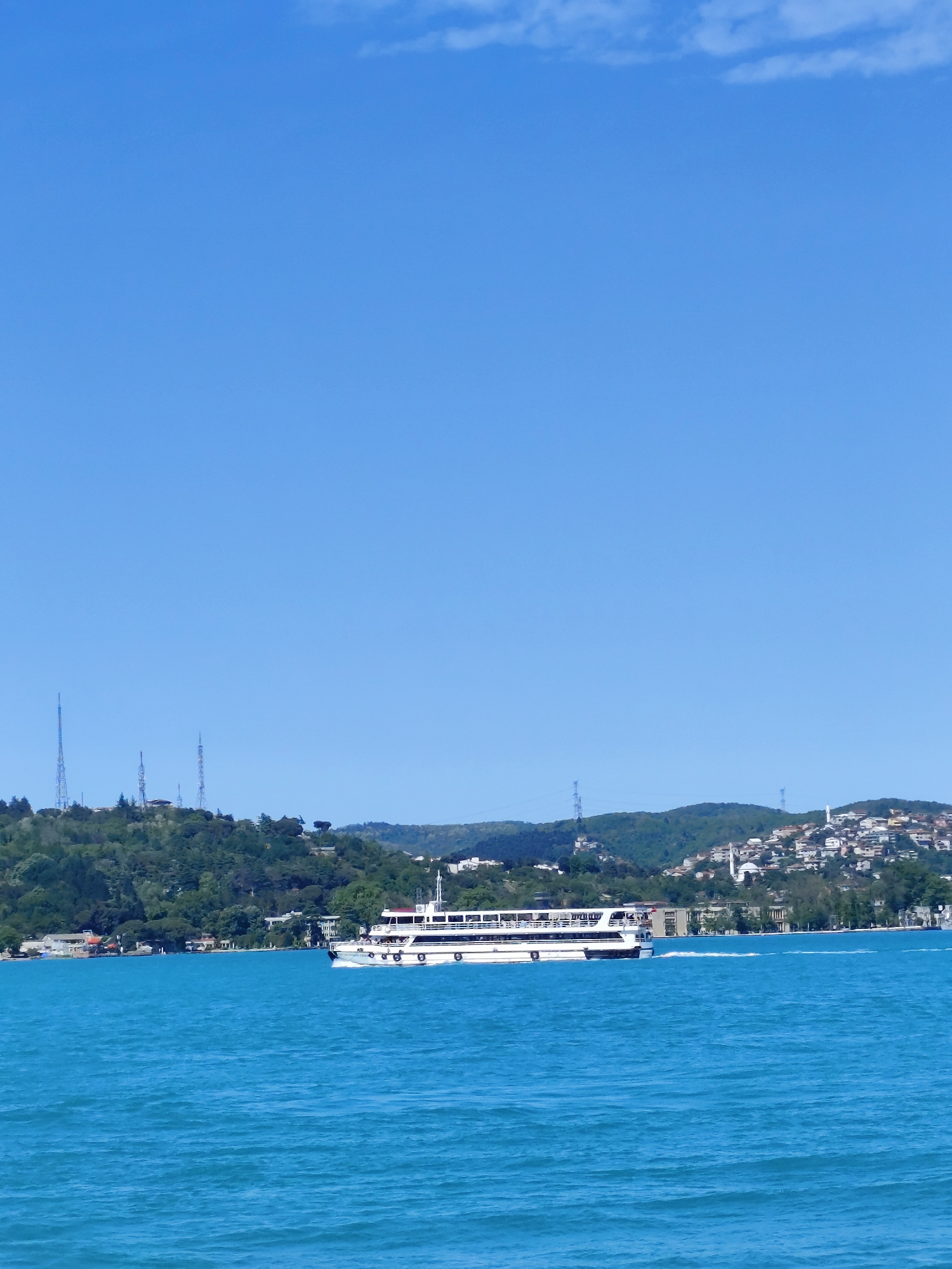 The image shows a white ferry moving across a body of water, likely the Bosphorus strait, in Sarıyer, Turkey. The ferry is seen against a backdrop of green hills and buildings, suggesting a coastal landscape. Several tall telecommunications towers can be seen on one of the hills. The water appears to be a clear, turquoise color under a clear blue sky with minimal clouds. The city of Sarıyer is discernible on the distant shoreline.