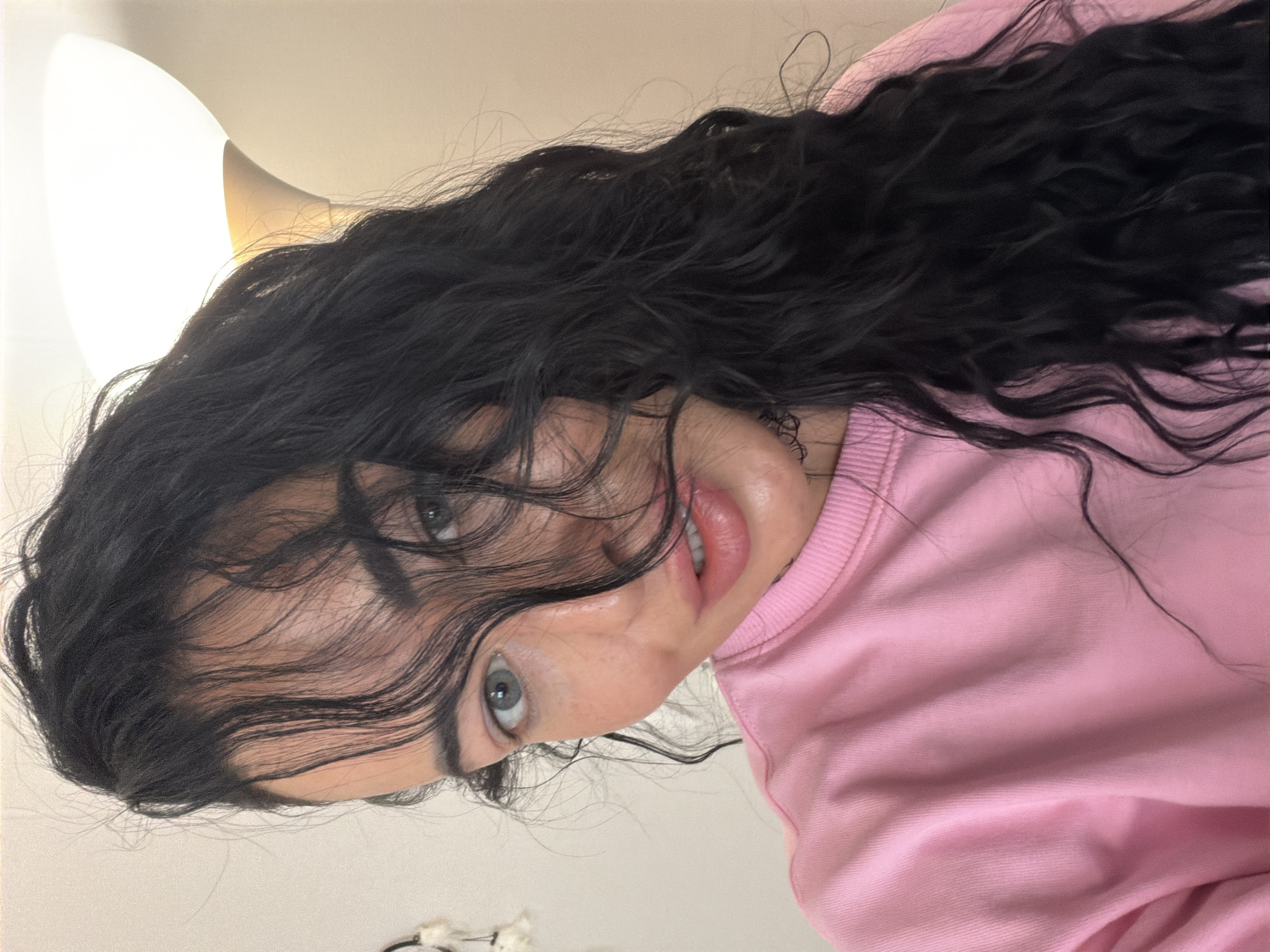 A medium close-up, low-angle shot depicts a woman with long, wavy black hair partially obscuring her face. Her skin is light, and she has visible eyes, eyelashes, eyebrows, and light pink lips. She is wearing a pink long-sleeved sweater. A lamp with a yellow glow is visible in the top left corner of the frame. The background is a light, neutral color, likely a wall or ceiling. No other people, objects, actions, or interactions are present in the image. The location in the city of Saint Petersburg, Russia is not possible to determine from this image.