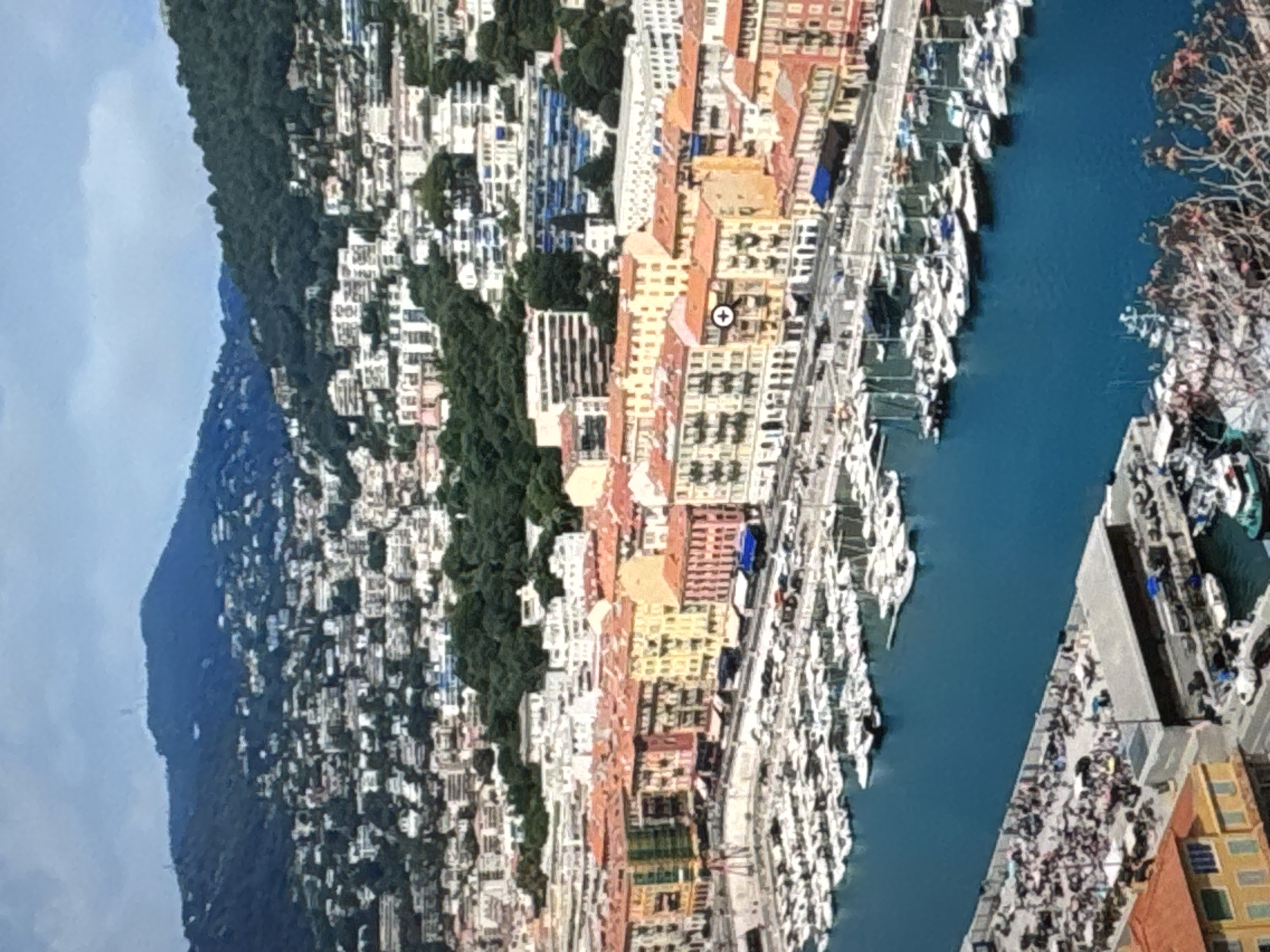 This image shows a panoramic view of a European coastal city, likely Nice, France, with a vibrant harbor filled with numerous boats. In the foreground, a bright blue body of water is densely packed with white sailboats and yachts. Along the waterfront, a promenade is lined with colorful buildings featuring red-tiled roofs and various architectural styles. Behind these buildings, the city climbs up a lush green hillside, densely populated with white and beige structures. The top of the image is dominated by a hazy sky and a large, dark green mountain range in the background, dotted with more buildings and some distant structures on its summit. There are no discernible individuals or specific actions taking place in the scene that would suggest it is located in Kaunas, Lithuania; the architecture and landscape are characteristic of the Mediterranean coast.