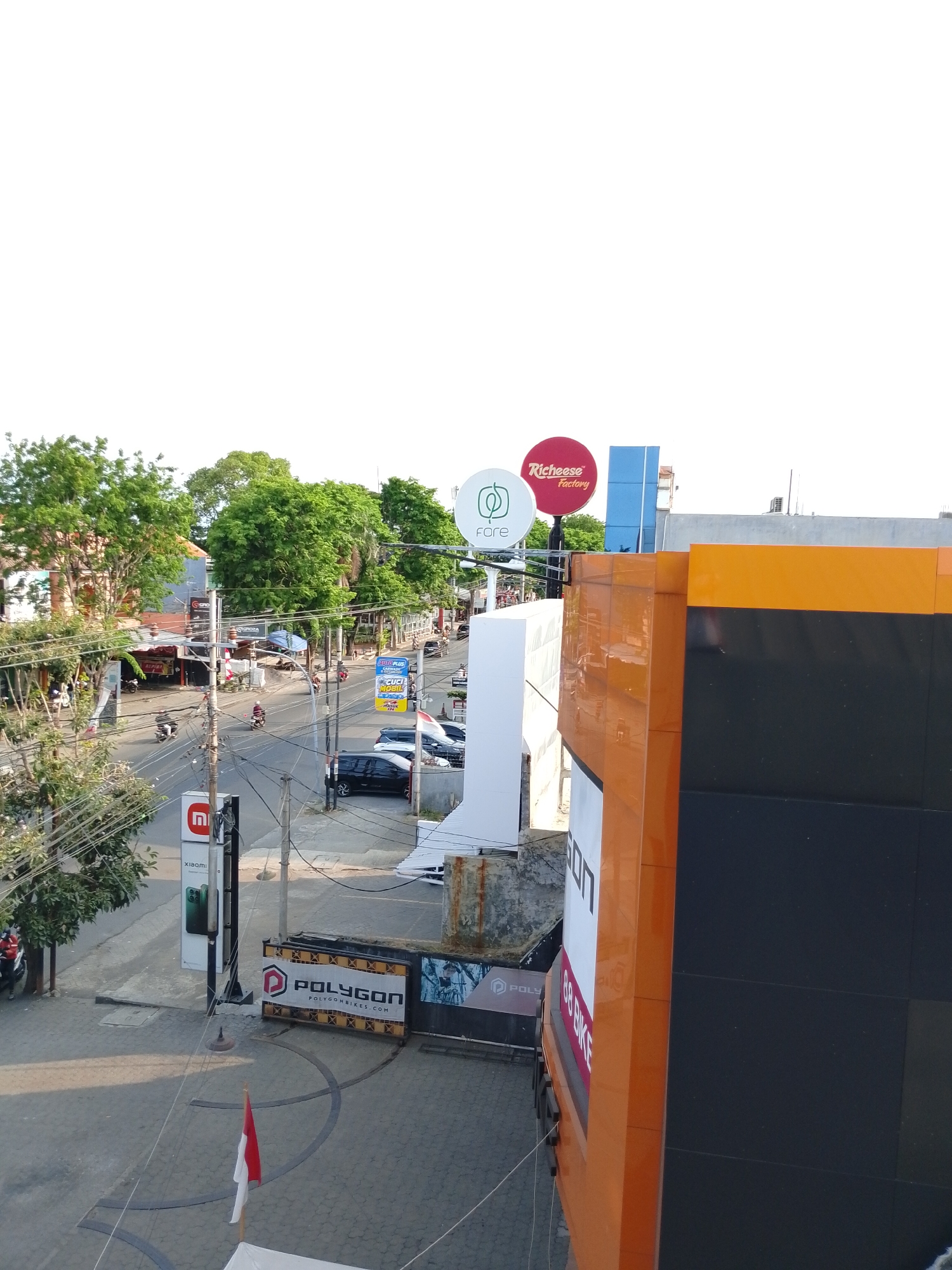 This is an eye-level, slightly elevated shot of a street scene in Pasuruan, Indonesia. The foreground is dominated by the side of a modern building with orange and black panels. A sign on this building, partially visible, reads "88 BIKE". To the left, a white structure is also visible.
Further down the street, a traffic intersection is captured. Cars and motorcycles are present on the road. A prominent sign for "Richeese Factory" is visible in red, along with a white circular sign that says "fare". Another sign with a red and white Indonesian flag is planted in the foreground. There are several trees lining the street, providing a green contrast to the urban environment. Overhead, a complex web of electrical wires is evident. The sky is a clear, bright white, suggesting daylight.