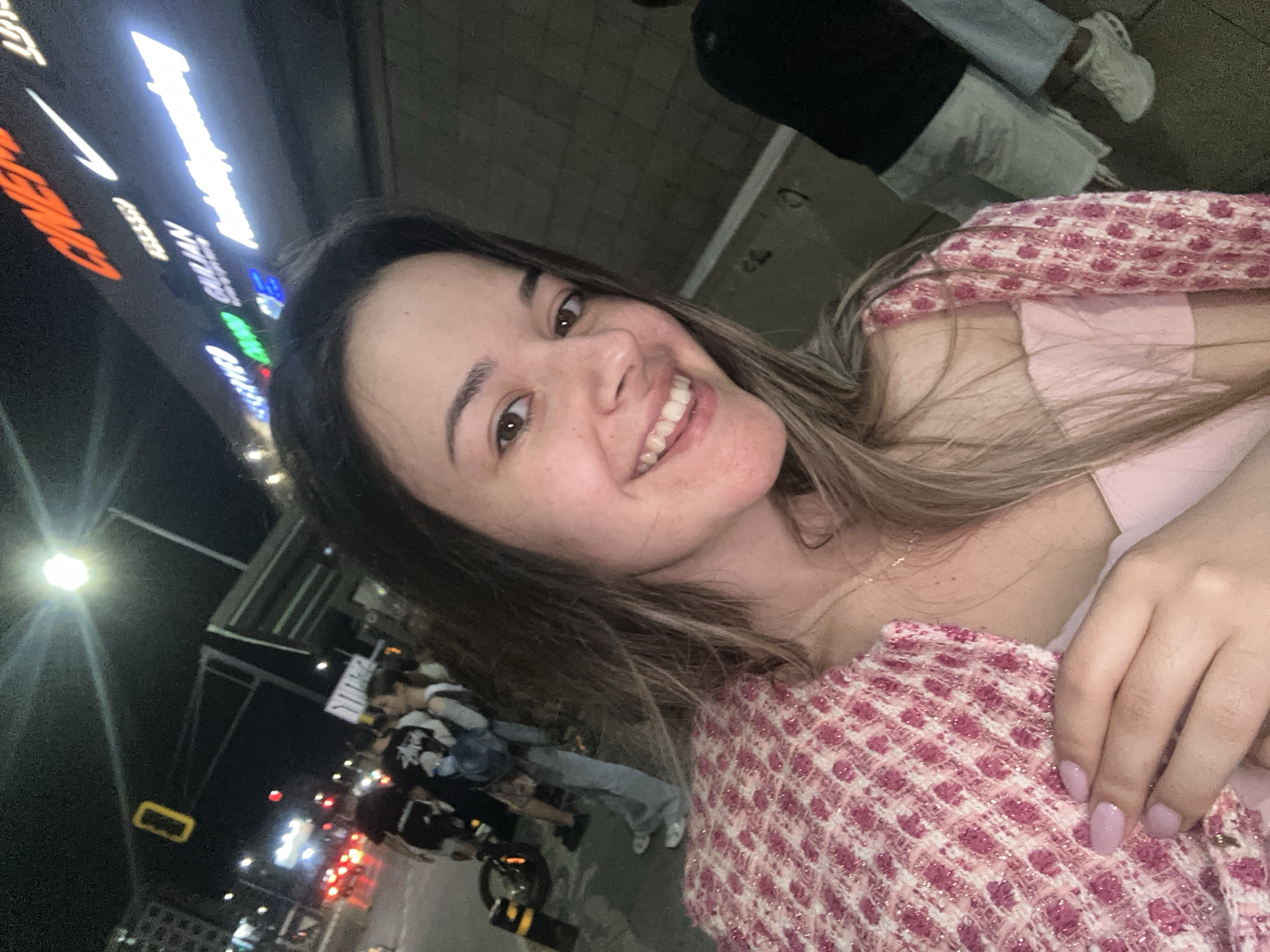 This is a nighttime photograph taken in an outdoor urban setting in Sofia, Bulgaria, specifically in the vicinity of Paradise Center.
In the foreground, a young woman with long, light brown hair and dark eyes smiles directly at the camera. She is wearing a light pink camisole and a textured pink and white jacket, possibly tweed, with visible glittery threads. Her left hand, adorned with light pink nail polish, is resting on her jacket.
The background reveals a street scene and a commercial building. To the left, a street is visible with blurred red taillights from moving vehicles. A bright street light casts illumination, and a yellow rectangular traffic light hangs overhead. Several individuals are gathered on a paved area, including one person wearing a white t-shirt with a "Stussy" logo and another with a light blue backpack. A motorcycle or scooter is partially visible, parked next to a black and yellow striped bollard.
Above and to the right of the woman, the illuminated signs of a building are prominent. Identifiable signs include "CINEMA" in orange lettering, "GAIJIN" in green, the white Nike swoosh logo, and "Lamborghini" in white lettering. Another white sign is partially visible to the right of "Lamborghini." To the lower right, a person in a black t-shirt and light shorts is partially visible, along with another individual wearing light blue jeans and white sneakers. The ground surface appears to be paved concrete.