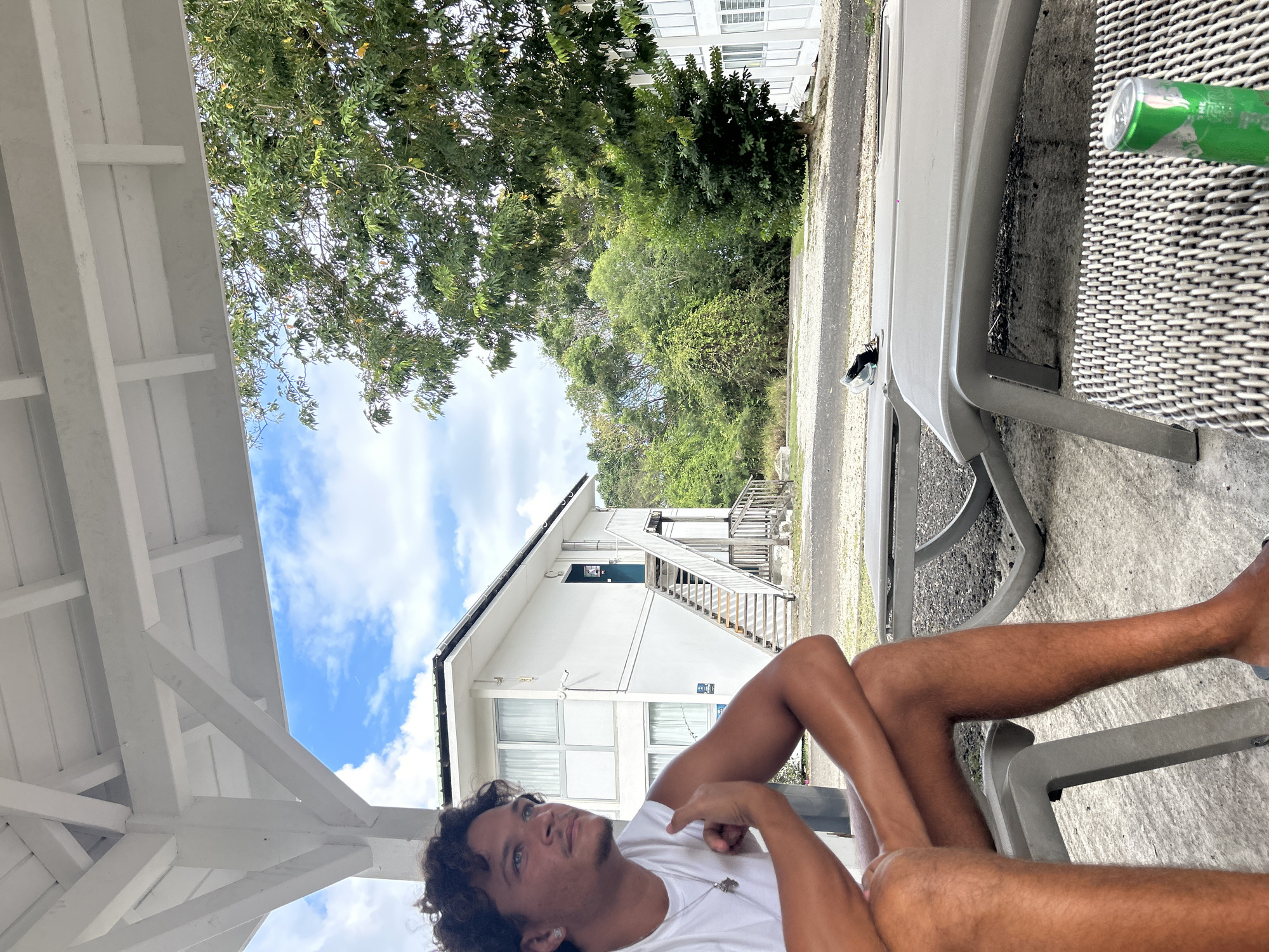 A man reclines on a grey plastic lawn chair under a white wooden porch in Sainte-Anne, France. The sky is partly cloudy and a white two-story building with an exterior staircase is visible. Green foliage is in the background. A green can sits on the table next to him.