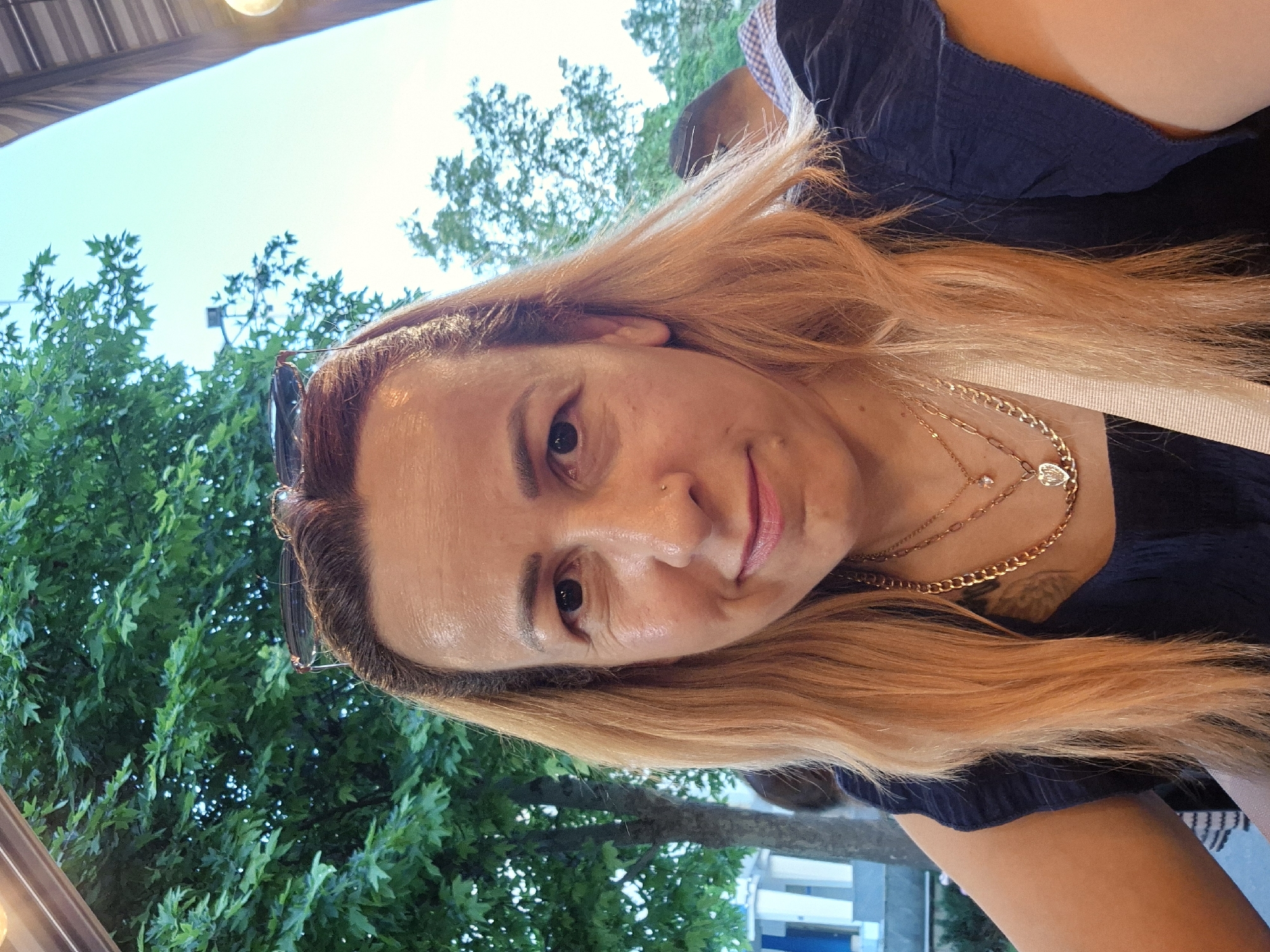 A medium shot captured from a slightly low angle shows a fair-skinned woman with light brown hair pulled back and sunglasses resting on her head. She is wearing a dark blue top with short, ruffled sleeves and multiple gold necklaces, one of which has a heart-shaped pendant. A light-colored strap crosses her chest. Her face is turned towards the camera, with her head tilted slightly to the left. She has a small nose ring and a mole on her left cheek. Behind her, green trees and a clear sky suggest an outdoor setting. The background also shows parts of buildings and another person, partially visible and out of focus. The lighting appears natural, indicating it is daytime. The image quality is good, with clear detail of the subject and her immediate surroundings.