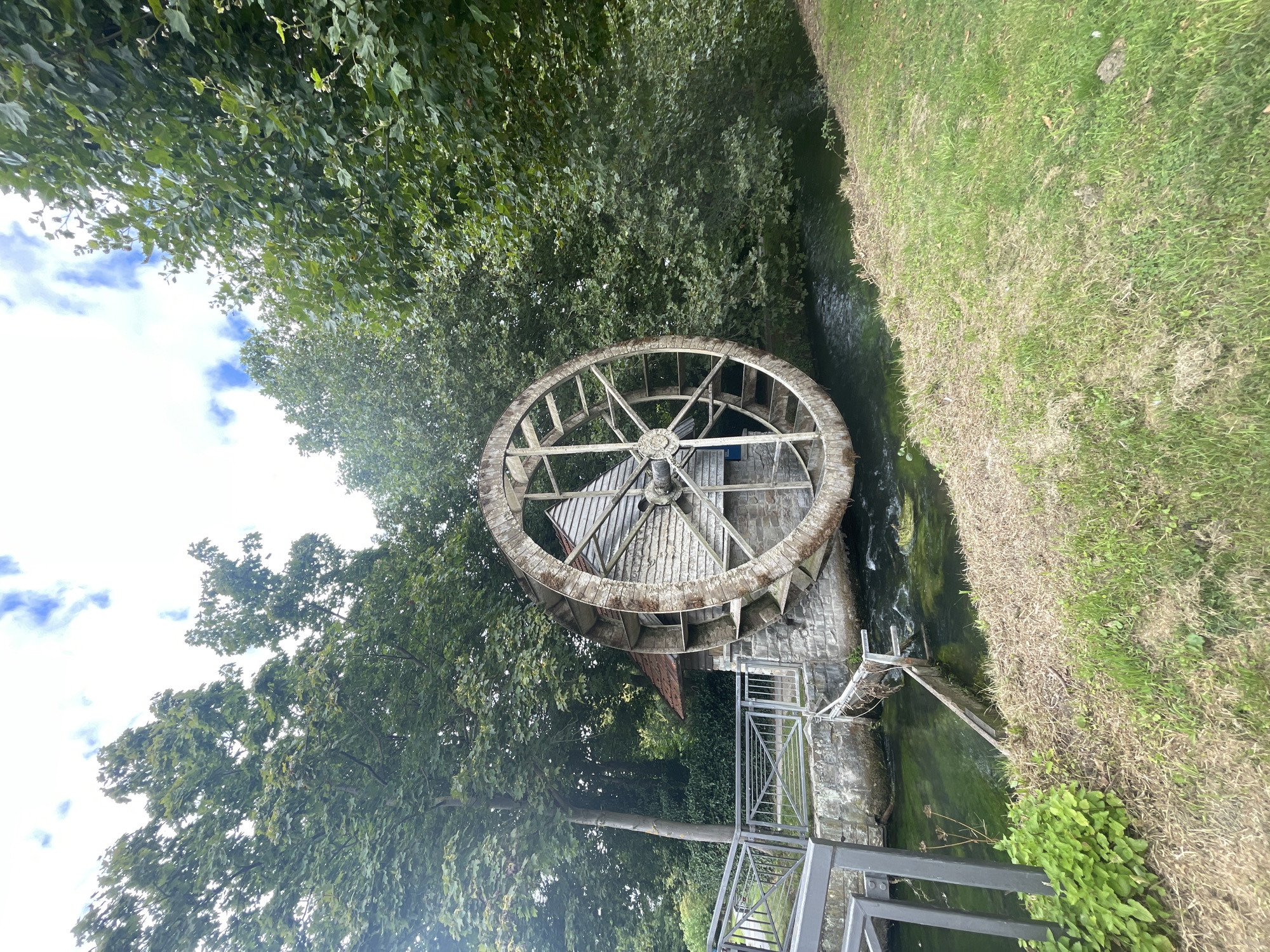 The image shows a large, wooden water wheel situated next to a stone building on the banks of a stream. The water wheel appears to be old and weathered, with visible spokes and buckets. The building has a red-tiled roof and is constructed from stone and wood. The stream is shallow and appears to be flowing gently. Surrounding the scene are lush green trees and grass, suggesting a natural or park-like setting. The sky above is partly cloudy. The image is taken from a slightly elevated angle, looking towards the water wheel and building. This scene is located in the city of Salzkotten, Germany, specifically at the historic watermill "Ueding Mühle." There are no people visible in the image.