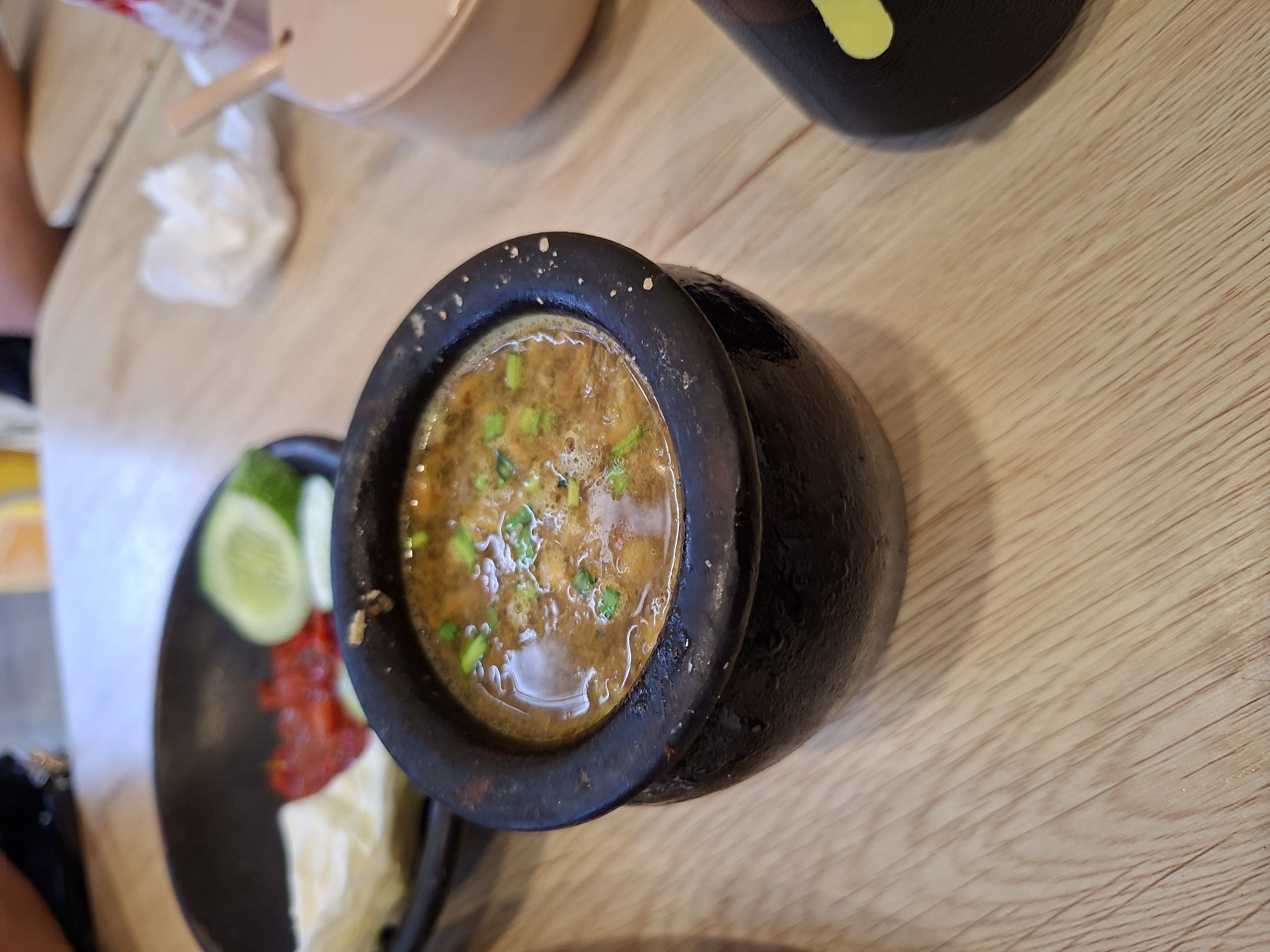 A close-up shot captures a small, dark ceramic pot filled with a simmering, brown soup, garnished with chopped green herbs. The pot sits on a light-colored wooden table, suggesting a dining setting. In the blurred background, a black plate holds what appears to be a portion of a meal, including slices of cucumber, a dollop of red sauce, and some white food item. To the right, a portion of a pink container is visible. The overall composition suggests a culinary scene, possibly at a restaurant or home, in Banjaratma, Indonesia.