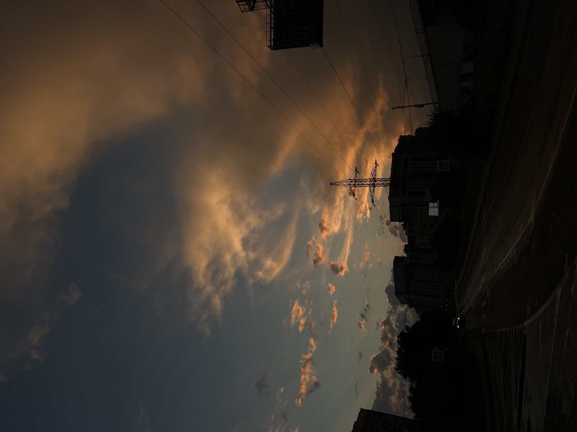 The image captures a sunset scene in Krasnodar, Russia. The sky is filled with dramatic clouds tinged with warm orange and yellow hues, contrasting with the dark, deep blue of the impending night. Several power lines stretch across the frame, supported by transmission towers, indicating an urban or semi-urban setting. Silhouetted buildings are visible on the right side of the image, their details obscured by the low light. A dark road curves into the distance, possibly wet from recent rain, reflecting the faint light of the sky. A pedestrian crossing sign is visible on the left, adding another cue to the urban environment. The overall mood is serene and perhaps a little melancholic, characteristic of a fading day. No people are visible, and the focus is on the interplay of light and shadow, the urban infrastructure, and the natural beauty of the sky at dusk.