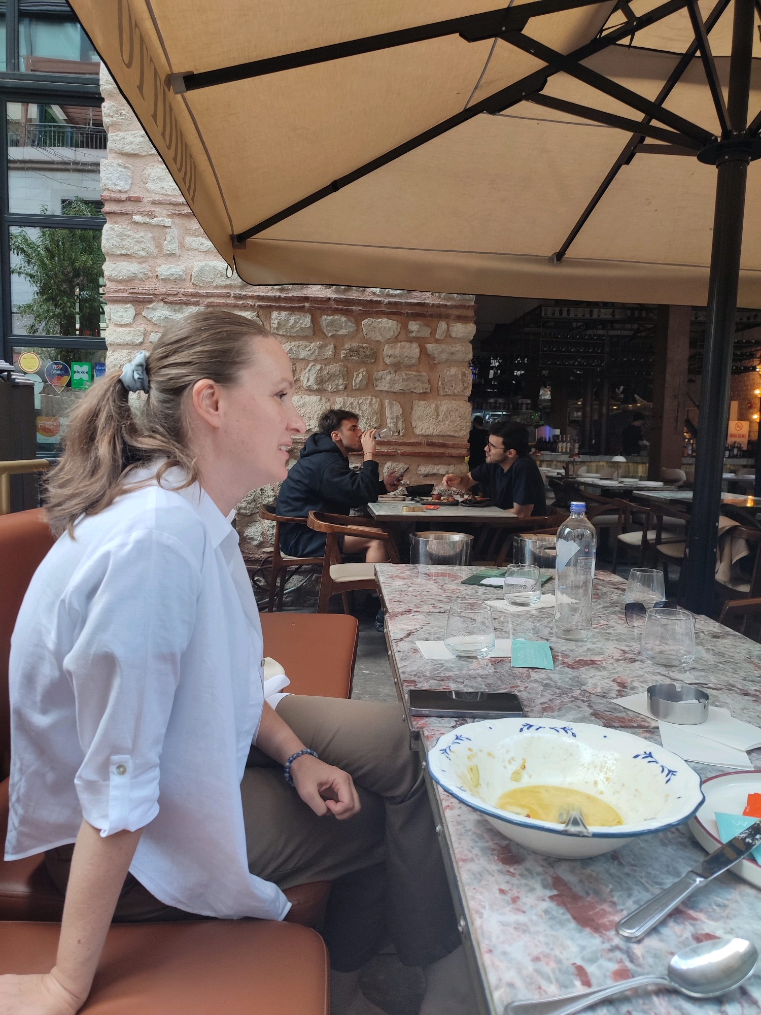 A woman with light brown hair pulled into a ponytail is seated at a marble-topped table, wearing a white button-down shirt and brown pants. She is looking to her right. In the background, two men are seated at another table. One man in a black hoodie is drinking from a glass. The other man, wearing glasses and a black t-shirt, gestures with his hand. The scene appears to be an outdoor seating area of a restaurant with a large beige umbrella providing shade. In the distance, a street with trees and buildings is visible, suggesting an urban environment. The woman is at a table with several glasses and a mostly empty bowl containing a yellow substance, possibly food residue. A smartphone is visible on the table. The architectural style of the building in the background, with its stone facade and red brick accents, may indicate a location in Zeytinburnu, Turkey.
