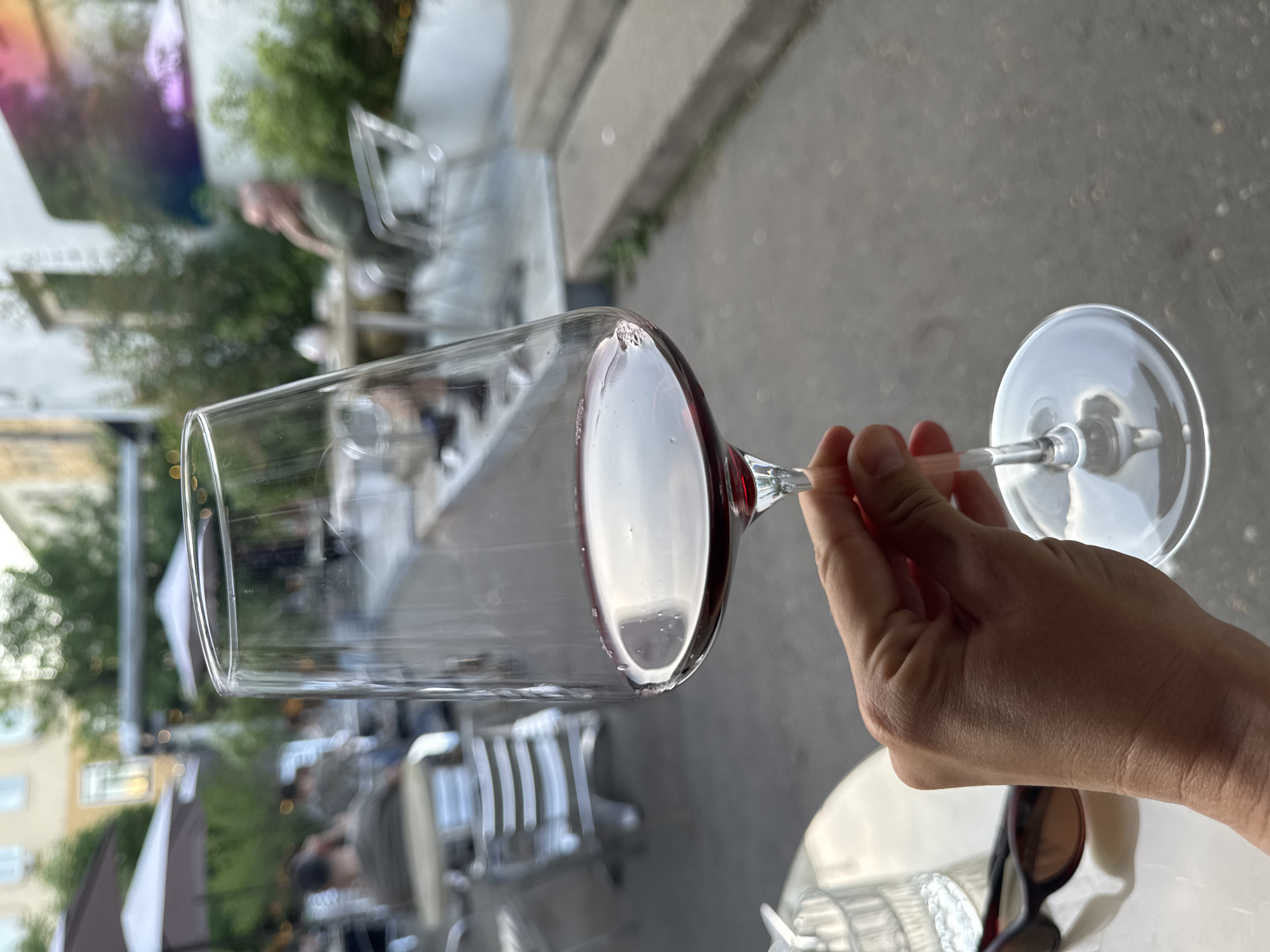 A hand holds a wine glass with red wine, tilted horizontally. The background shows an outdoor seating area with metal chairs and tables, trees, and buildings. The scene may be located in Moscow, Russia.