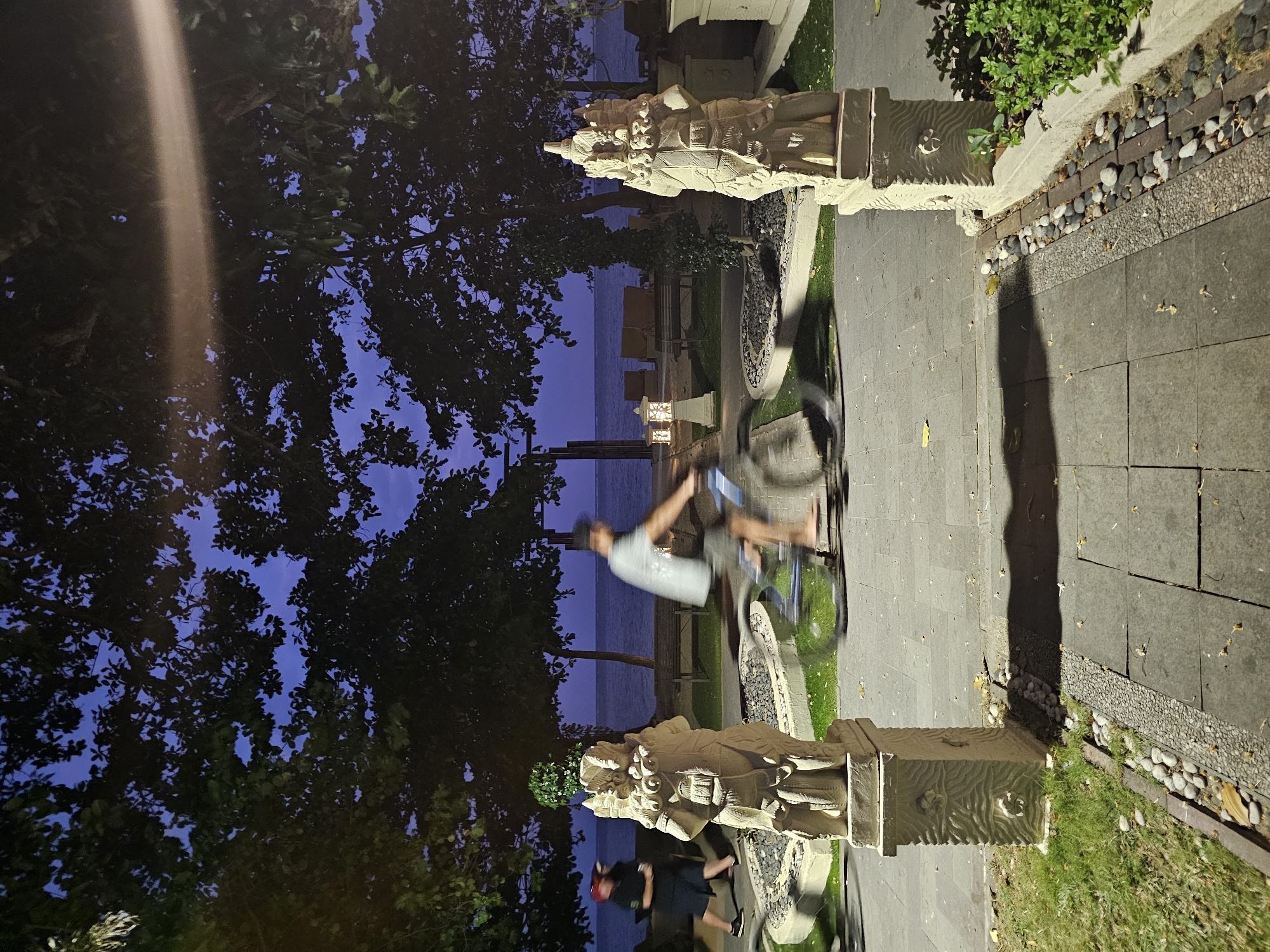 A person is riding a bicycle on a paved pathway next to the ocean at dusk. The sky is a deep blue, indicating the sun has set, and the water is calm. Two large, ornate stone statues, possibly Balinese in style, flank the pathway. The statue on the left is partially obscured by the dark leaves of a large tree. The statue on the right stands beside a well-maintained garden bed with grass and decorative stones.
The cyclist, seen in motion blur, is wearing a light-colored t-shirt and shorts, and a hat. Their face is not clearly visible due to the motion. Another person, also blurred and wearing a red helmet, is visible in the lower left portion of the image, seemingly walking or standing near the base of the left statue.
The pathway is made of large, grey stone tiles, with a border of smaller, darker stones and gravel. Fallen leaves are scattered on the pathway and grass. A small, traditional lantern with a lit bulb is visible near the water's edge, casting a warm glow. The overall atmosphere suggests a peaceful evening by the sea, with hints of local culture due to the statues. The location cues, such as the statues and the tropical foliage, along with the "Denpasar, Indonesia" context, suggest this scene is likely in Bali.