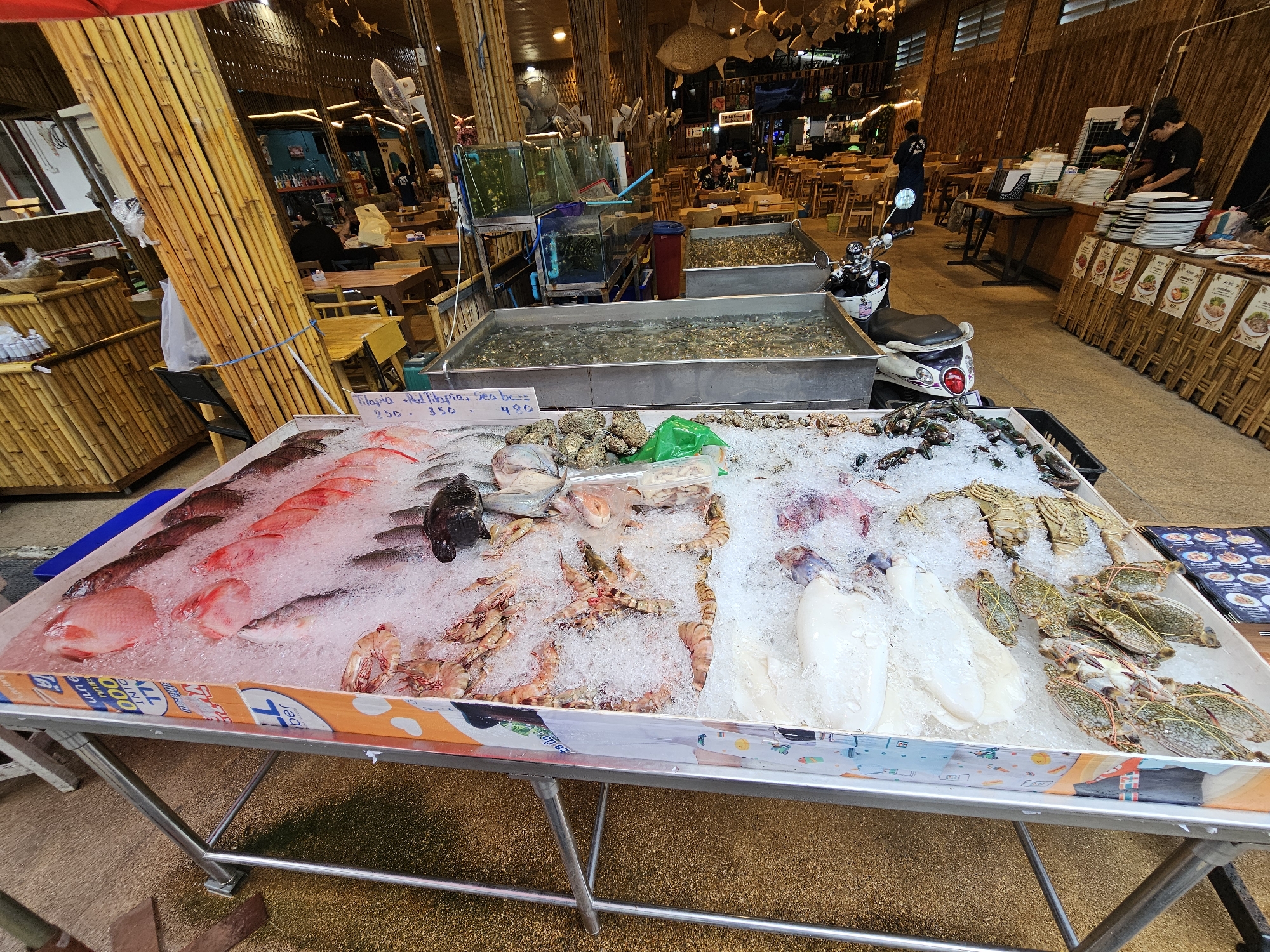 In an open-air restaurant in Bangkok, Thailand, a display of fresh seafood is arranged on a metal-framed table covered with ice. The seafood includes various fish, shrimp, squid, crabs, and lobsters, each meticulously laid out. Behind the seafood display, large tanks filled with live shrimp are visible, alongside a motorcycle parked nearby. The restaurant's ambiance is rustic, with bamboo accents and traditional Thai decor. Patrons are seated at wooden tables in the background, enjoying their meals. The scene captures a bustling marketplace where fresh seafood is prepared and served, reflecting the vibrant culinary culture of Bangkok.
