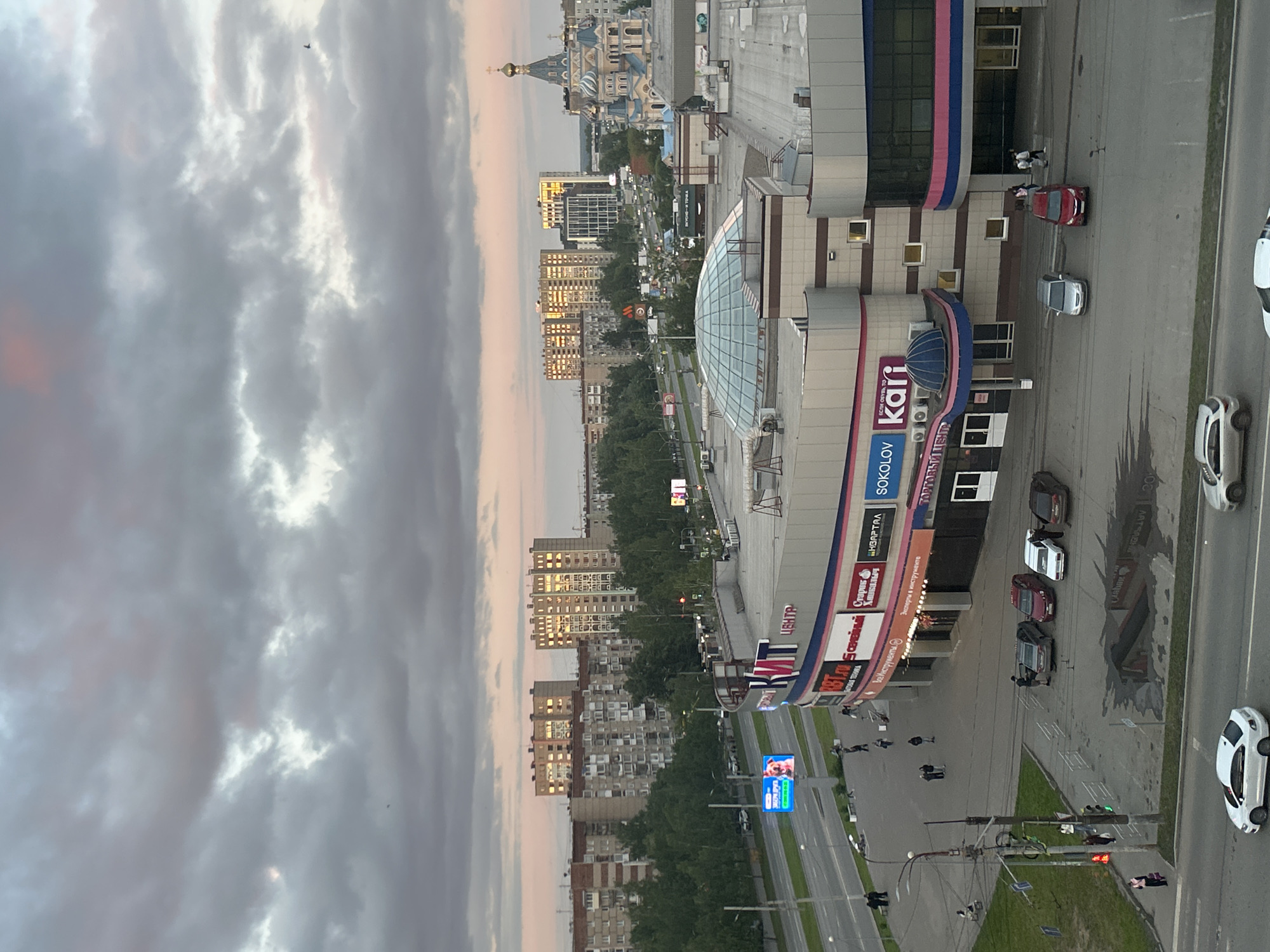 This media file presents an elevated view of an urban scene in Izhevsk, Russia, captured during what appears to be the late afternoon or early evening. The sky is largely overcast with grey clouds, but soft pink and orange light breaks through, illuminating parts of the clouds and suggesting a transitional time of day. A small, dark object, possibly a bird or drone, is visible against the clouds in the upper left.
In the foreground and mid-ground, a prominent multi-story shopping center dominates the right side of the frame. It features a modern design with sections of blue, pink, and grey paneling, and a distinctive glass-domed entrance. Several cars are parked in front of the center, and a few pedestrians are visible on the sidewalk. Notable visible text on the shopping center's facade includes large signs for "Kari" and "SOKOLOV," along with "ТОРГОВЫЙ ЦЕНТР" (Shopping Center), "ВСЕ ИНСТРУМЕНТЫ" (All Tools), and "Эксперты в инструменте" (Experts in tools). A slogan "ЕСЛИ ОБУВЬ, ТО..." (If shoes, then...) is also visible.
A wide, multi-lane road runs horizontally through the center of the image, with cars driving and parked along its edges. Traffic lights, streetlights, and a large blue LED advertising screen displaying dynamic content are also present. On the left side of the road, various multi-story residential and commercial buildings rise, many with windows reflecting the soft light. In the distant background, a grand and ornate church or cathedral with golden domes stands out against the horizon, characteristic of Russian architecture. Green trees are interspersed throughout the urban landscape. The scene depicts typical city life with a mix of commerce, residential areas, and historical landmarks.