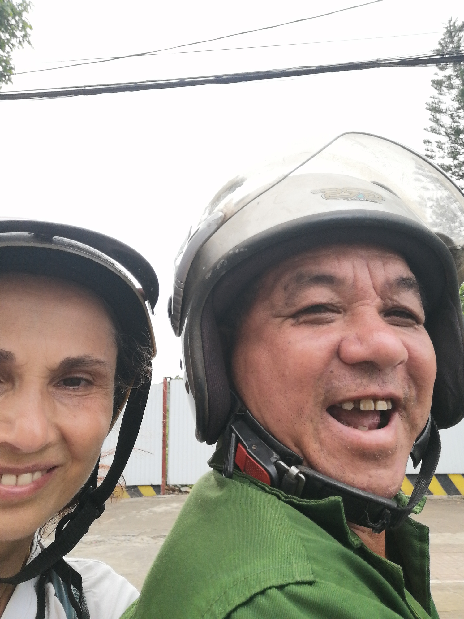 Two individuals, a woman and a man, are captured in a close-up, slightly high-angle selfie-style photograph. Both are wearing motorcycle helmets, suggesting they are on or near a motorbike or scooter. The woman is positioned on the left, partially visible, smiling subtly with dark hair framing her face and a white top. The man, on the right, is more prominently featured, wearing a green uniform-like shirt and smiling broadly with his mouth open, revealing his teeth. His helmet has a sticker with what appears to be a "2SP" or similar logo on the visor.
The setting appears to be outdoors during bright daytime, possibly under an overcast sky, as the sky in the background is mostly white. Visible in the background are power lines or communication cables strung across the upper frame, and some leafy tree branches on the left. Behind the individuals, there are industrial or commercial elements, including a white corrugated wall and a yellow and black striped barrier, possibly indicating a construction site or an industrial area. Both individuals exhibit expressions of happiness and enjoyment. The man's helmet has a strap with a red accent near his chin. Given the context, the photo is taken in Lao Cai Ward, Vietnam.
