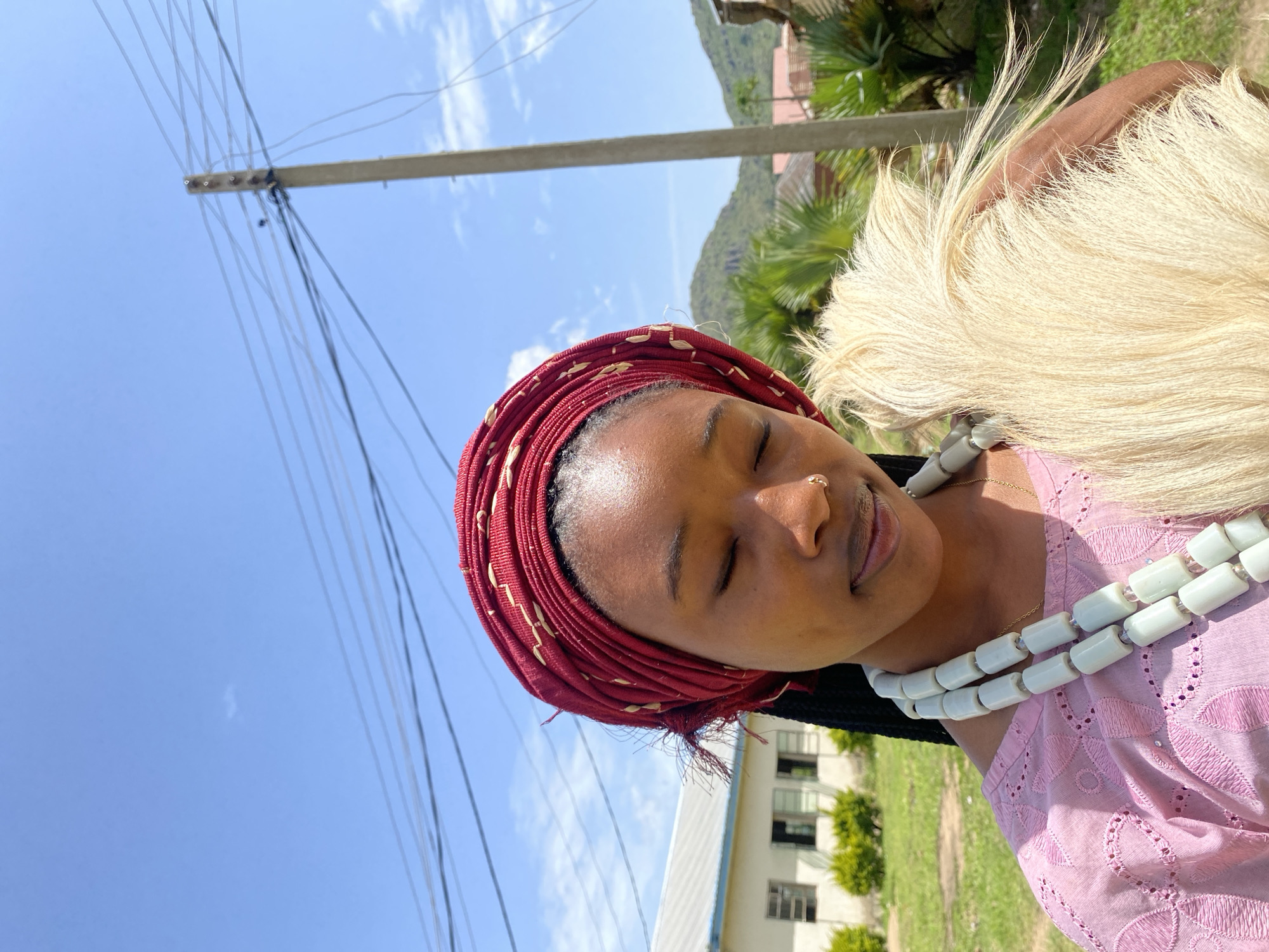 A young woman is captured in a close-up, slightly tilted, outdoor portrait. She is wearing a vibrant red headscarf with a gold woven pattern and a pink top adorned with intricate white embroidery. A striking, multi-strand necklace made of white, cylindrical beads is visible around her neck. Her eyes are closed, and her expression suggests a moment of peaceful contemplation or a pleasant pose for the photograph. A large, pale blonde wig or hairpiece is positioned behind her head, adding a dramatic element to her appearance.
The background reveals a clear blue sky with a few wispy clouds, indicating fair weather. To the left, a tall utility pole with numerous hanging wires dominates the scene, suggesting an urban or semi-urban environment. In the mid-ground and background, there are glimpses of buildings and lush green vegetation, including palm trees, hinting at a warm climate. The overall lighting suggests it is daytime, likely late morning or early afternoon, with bright sunlight casting soft shadows.
The setting, identified as Vom, Nigeria, provides a regional context for the woman's attire and the surrounding environment. The image conveys a sense of natural beauty and cultural richness, with the woman appearing serene and perhaps dressed for a special occasion. No visible text is present in the image.