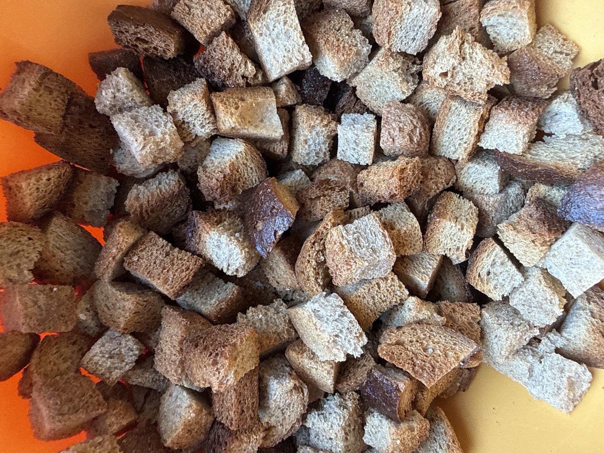 The image shows a close-up, overhead shot of croutons in an orange bowl or container. The croutons are roughly cube-shaped, varying in color from light tan to dark brown. They appear to be homemade or artisan-style due to their irregular shapes and varying textures. The background is a solid orange color. The image provides no information to confirm the location in Moscow, Russia.
