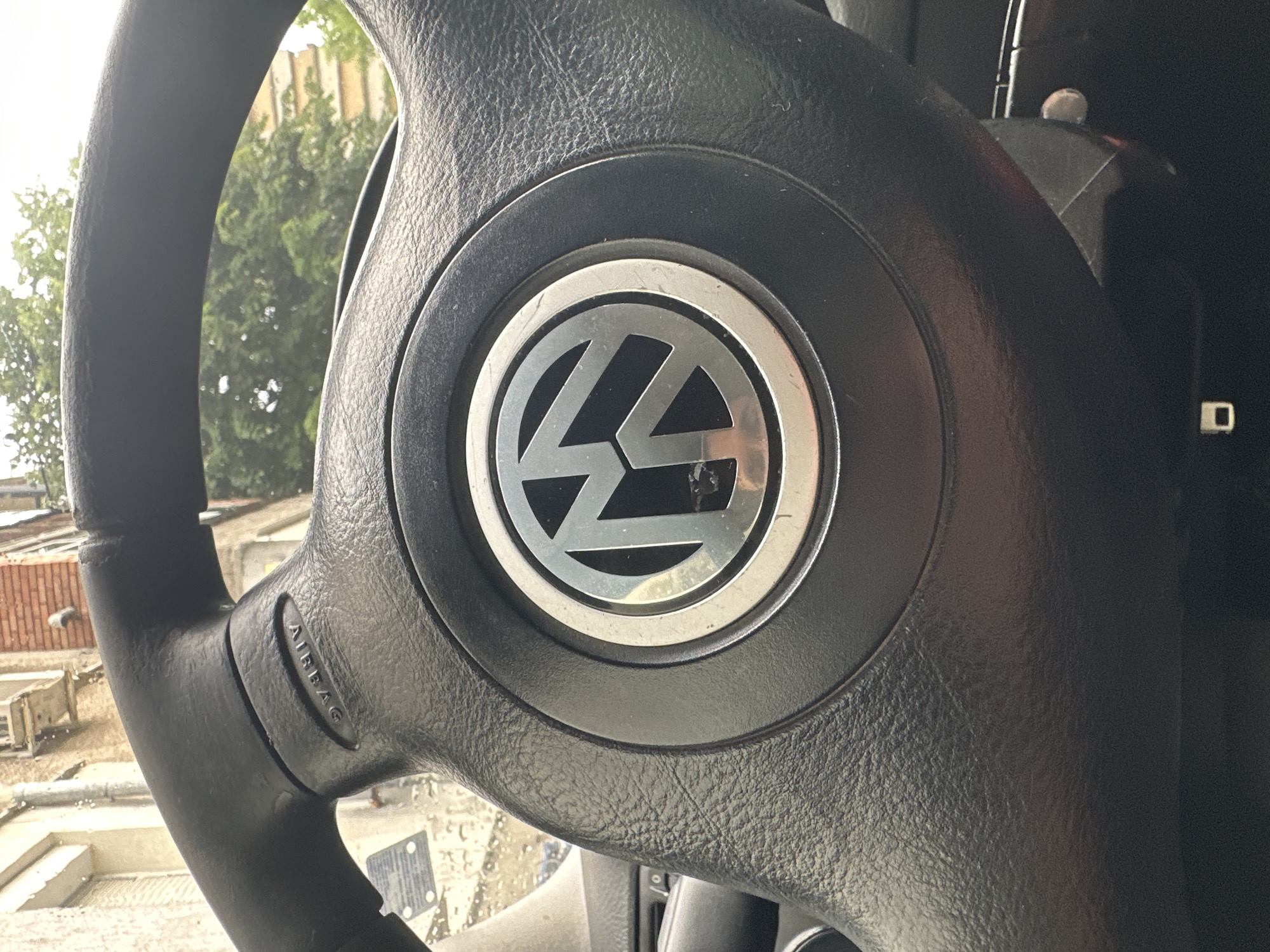 The image presents a close-up, angled view of a car's black steering wheel, seen from an interior perspective. The primary subject is the steering wheel, made of a textured, possibly synthetic leather or plastic material, with visible signs of wear. Centrally positioned on the steering wheel's hub is a prominent metallic silver Volkswagen (VW) logo, which exhibits some scratches and scuff marks, suggesting regular use.
On the left spoke of the steering wheel, the word "AIRBAG" is visibly embossed. To the right, a portion of the car's interior, dark and indistinct, can be seen.
Through the gaps in the steering wheel, the background reveals an outdoor setting during what appears to be daytime, indicated by bright, diffuse light. Lush green foliage from trees is visible, suggesting a natural element in the surroundings. Faint architectural elements, including a brick wall and other building structures, are discernible behind the greenery, hinting at an urban or suburban environment. No individuals are present, and no specific activity is taking place within the frame; the scene is static. The context provided places this vehicle in Radnevo, Bulgaria.