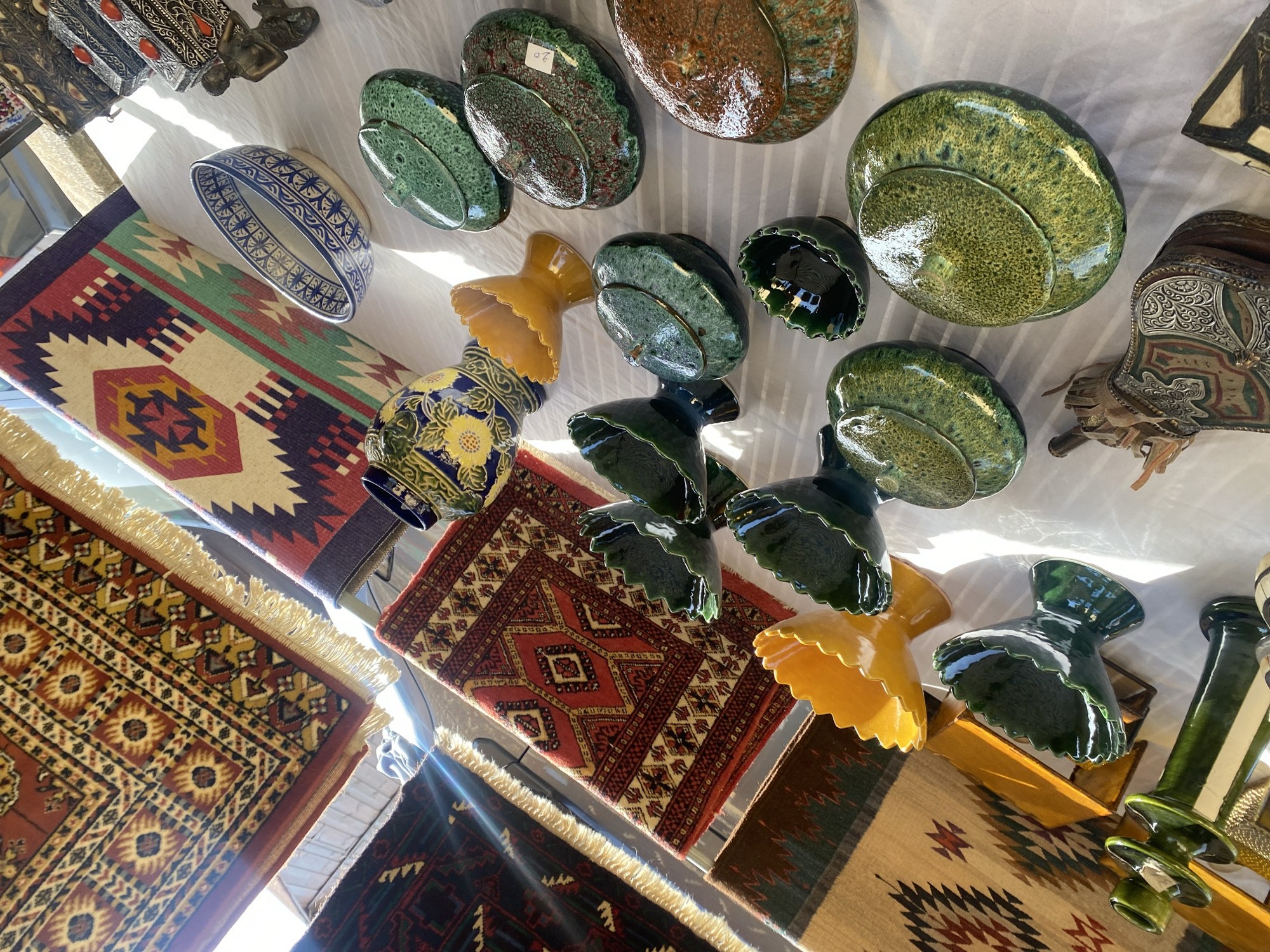The image depicts an outdoor market stall in Manilva, Spain, displaying a variety of handcrafted items. The foreground is dominated by several glazed ceramic bowls and vases in various shades of green, yellow, and brown, many with intricate, textured finishes. A decorative yellow bowl with a ruffled edge is prominently placed, alongside darker green, leaf-shaped vessels. Further back, a large blue and white patterned bowl sits near a colorful, geometric-patterned rug. Several other woven rugs and textiles with traditional designs are visible, some illuminated by sunlight streaming from the left. A dark blue ceramic vase with yellow floral motifs is also present. The stall appears to be set up on a white cloth or tablecloth, and elements of the surrounding environment, such as sunlight and shadows, suggest an outdoor setting. The overall impression is one of a vibrant display of artisanal goods.