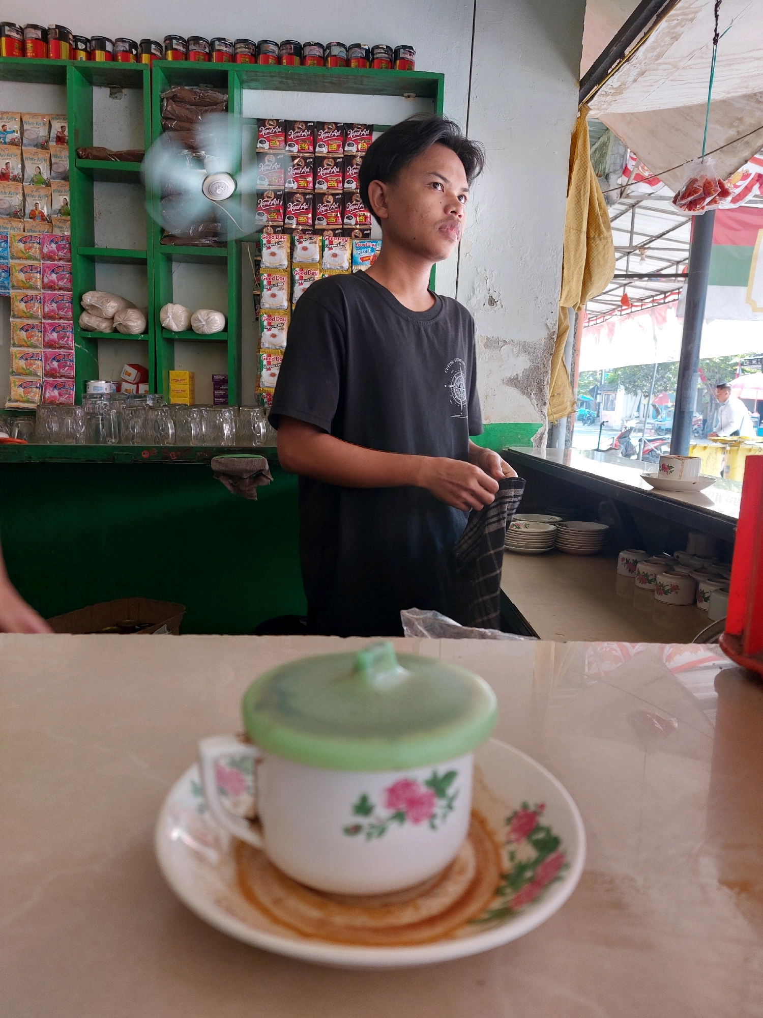 In Sumuragung, Indonesia, a young man stands behind a counter in what appears to be a small shop or food stall. He wears a dark t-shirt and is looking off to the right, holding a checkered cloth. The counter is cluttered with various items, including stacks of saucers, cups, and packets of instant coffee. Behind him, shelves are filled with more coffee packets and other goods. A small fan is mounted on the wall, its blades blurred with motion. In the foreground, a small cup with a green lid sits on a saucer, also decorated with floral patterns. The overall impression is of a modest, bustling local establishment.