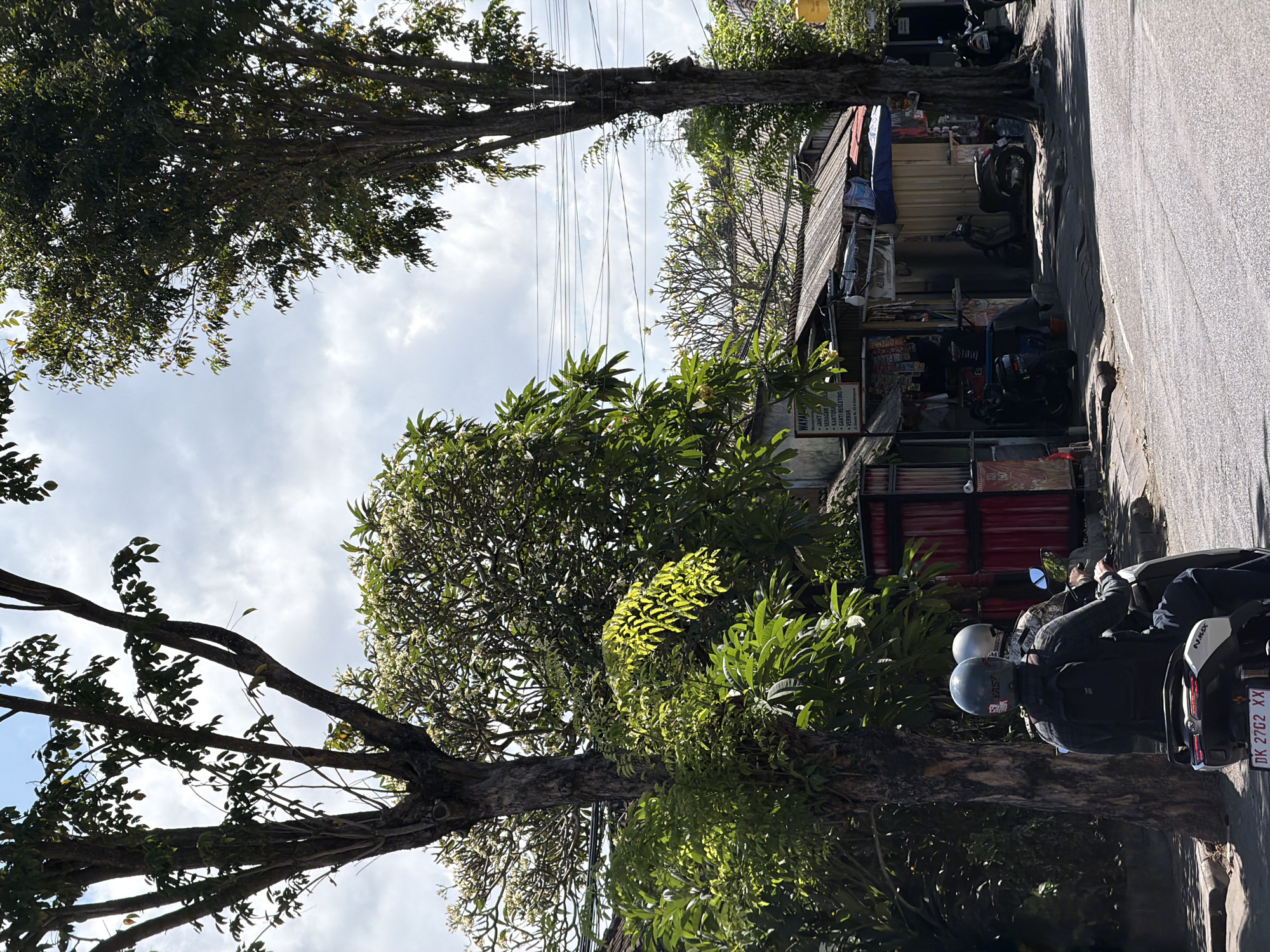 A street scene in Kuta, Indonesia, features two people riding a motorcycle in the foreground, heading away from the viewer. The driver wears a dark jacket and a grey helmet, while the passenger wears a camouflage jacket and a white helmet. The motorcycle is white with black accents and a visible license plate reading "DK 2702 XX."
To the right of the motorcycle, a row of small storefronts with corrugated metal roofs lines the street. One storefront has a sign with text in Indonesian, including the words "JAHIT," "SERAGAM," "KANTOR," and "VERMAX." Several other motorcycles are parked in front of these shops.
Lush green trees with dense foliage and some white flowers are prevalent throughout the scene, some casting shadows on the road. A large, tall tree stands prominently on the right side of the image. Overhead, power lines crisscross against a bright sky with scattered clouds. The road is asphalt and appears to be a typical street in a tropical urban environment. The overall impression is one of a sunny day in a bustling Indonesian town.