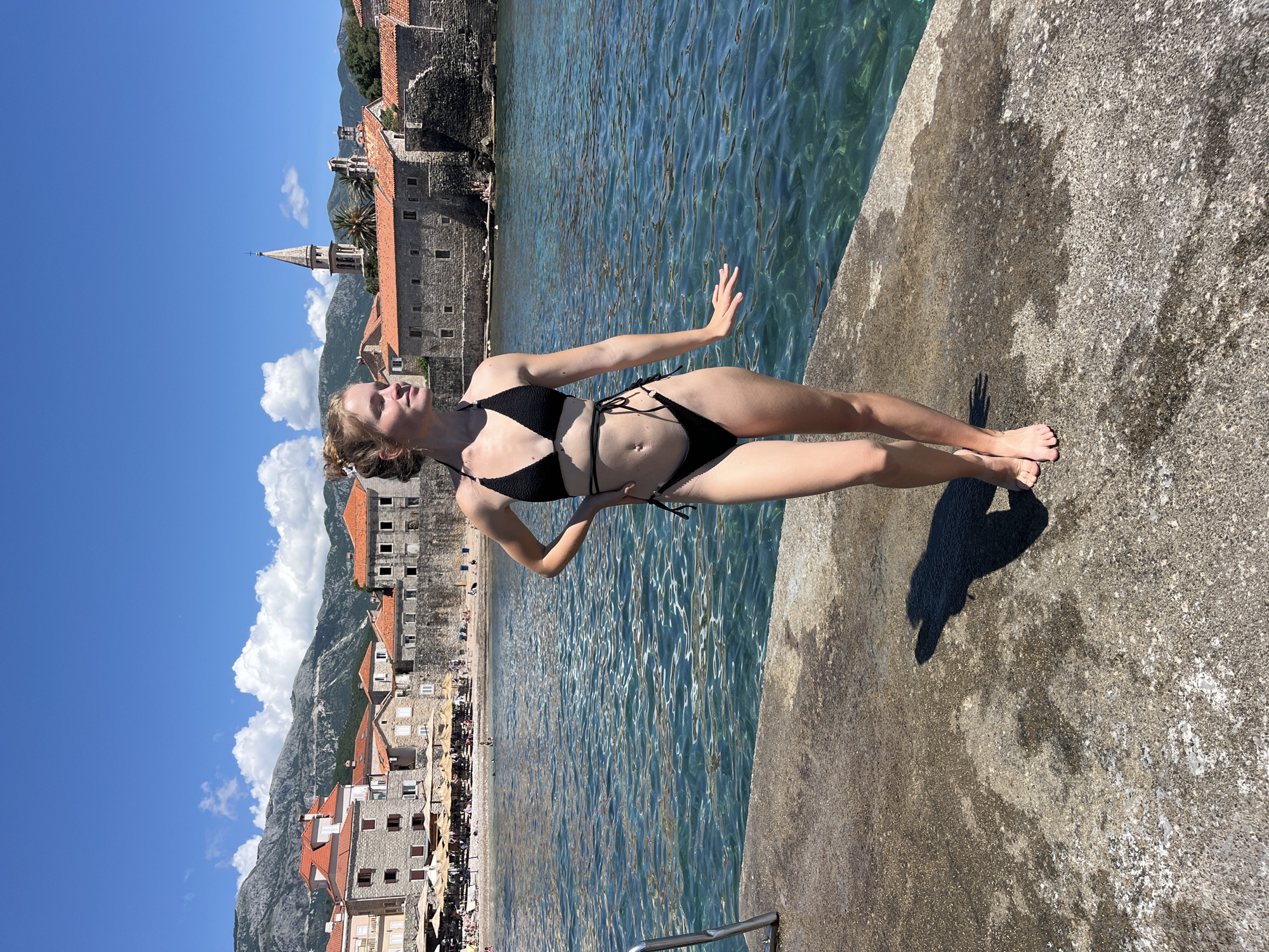 This image depicts a vibrant, sunny day in Budva, Montenegro, with a young woman posing on a concrete pier or wall beside the sea. The woman, with light-colored hair tied up, wears a black string bikini with a textured top. She is barefoot and strikes a pose, her head tilted slightly back with eyes closed or looking up towards the sun, her right arm extended, and her left hand on her hip. Her shadow is cast distinctly on the sun-drenched concrete.
To the right of the woman, the clear, blue-green sea extends, showing sunlit ripples on its surface. In the background, a picturesque old town with ancient stone buildings and traditional red-tiled roofs is visible. A prominent church steeple with a pointed spire stands tall within the town's architecture. Majestic mountains rise behind the town under a bright blue sky adorned with white cumulus clouds.
The scene suggests a leisurely, mid-day vacation moment, indicated by the strong sunlight and clear skies, ideal for enjoying the beach and sea. In the distance, a sandy beach with numerous umbrellas and people can be seen, highlighting the area's popularity. A metal handrail or ladder partially visible at the bottom of the frame suggests entry into the water. The overall atmosphere is bright, serene, and inviting. No visible text is present in the image.